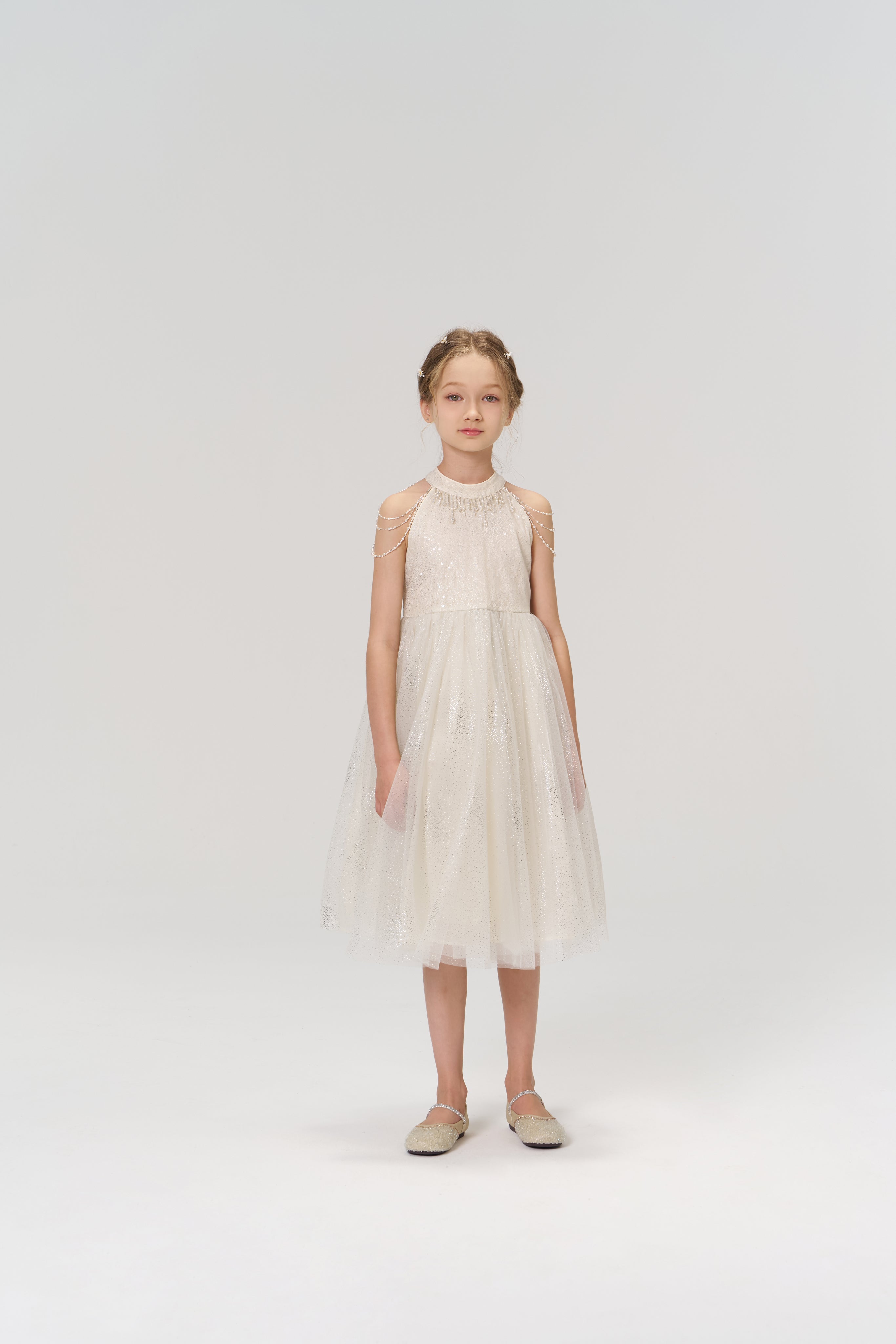 A young girl stands facing forward in the Gilded Whisper Dress by KIKI and JOJO—a beige tulle dress with sheer details—paired with flat shoes, set against a simple, light background.