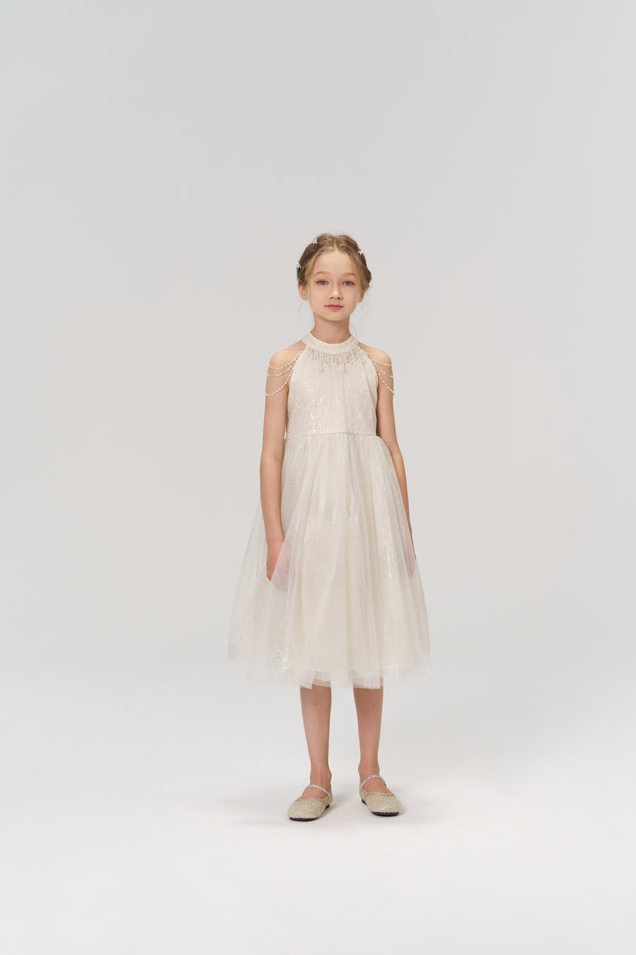 A young girl stands facing forward in the Gilded Whisper Dress by KIKI and JOJO—a beige tulle dress with sheer details—paired with flat shoes, set against a simple, light background.