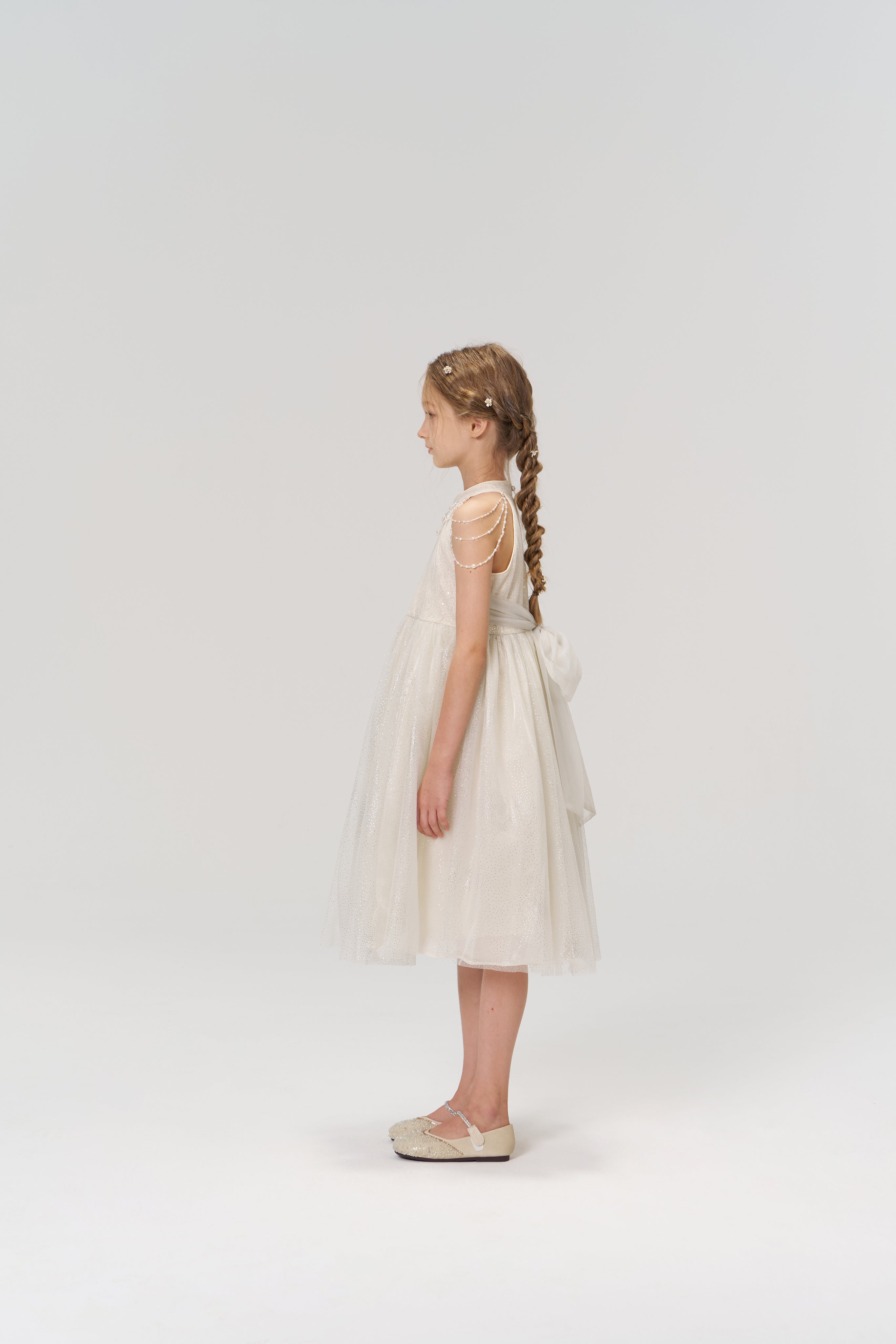 A young girl with braided hair stands in profile wearing the KIKI and JOJO Gilded Whisper Dress—a beige tulle dress with decorative shoulder straps and bow—paired with flat shoes, set against a plain light background.