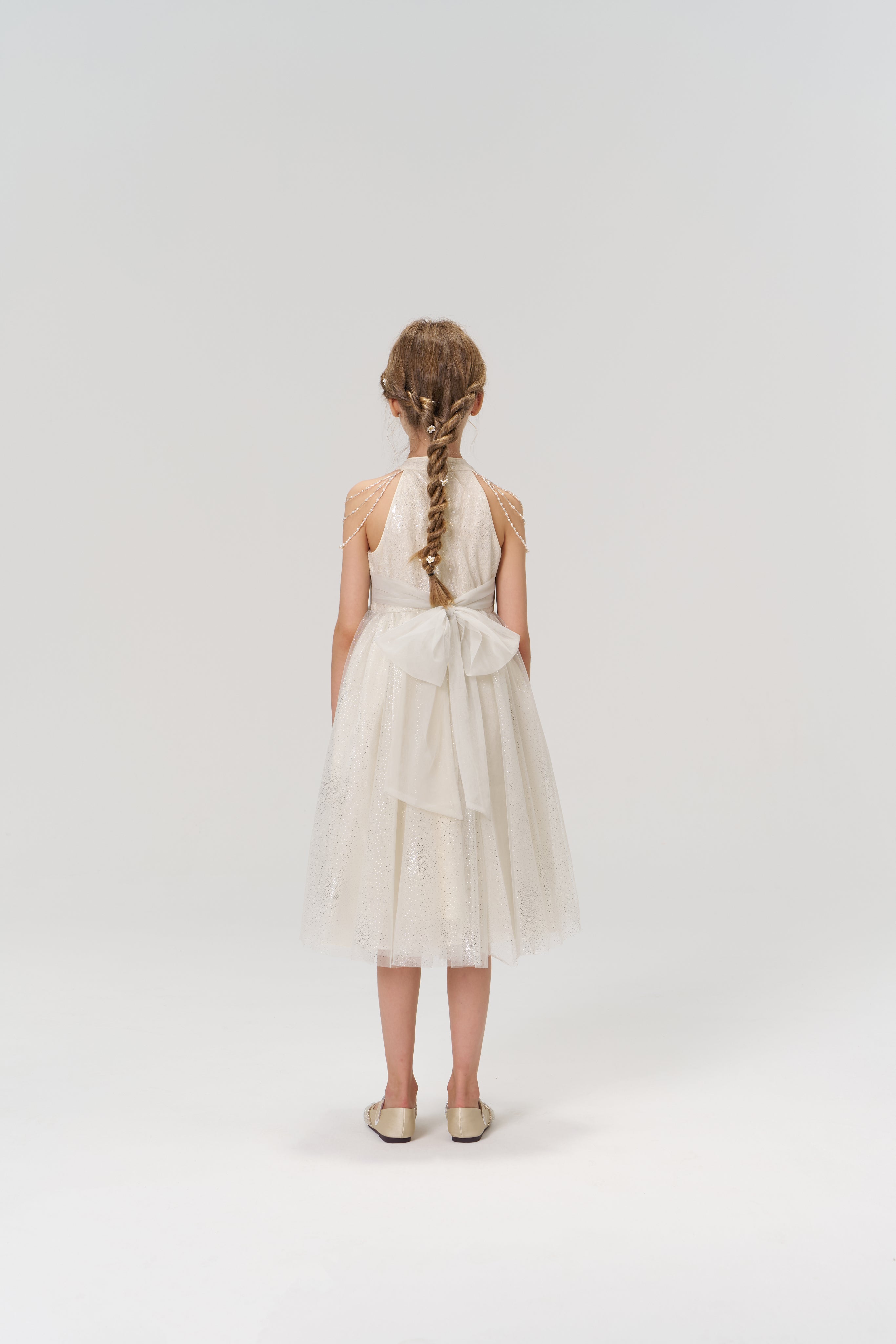 A young girl with braided hair, wearing the Gilded Whisper Dress by KIKI and JOJO—with a large back bow—stands facing away against a plain light background.