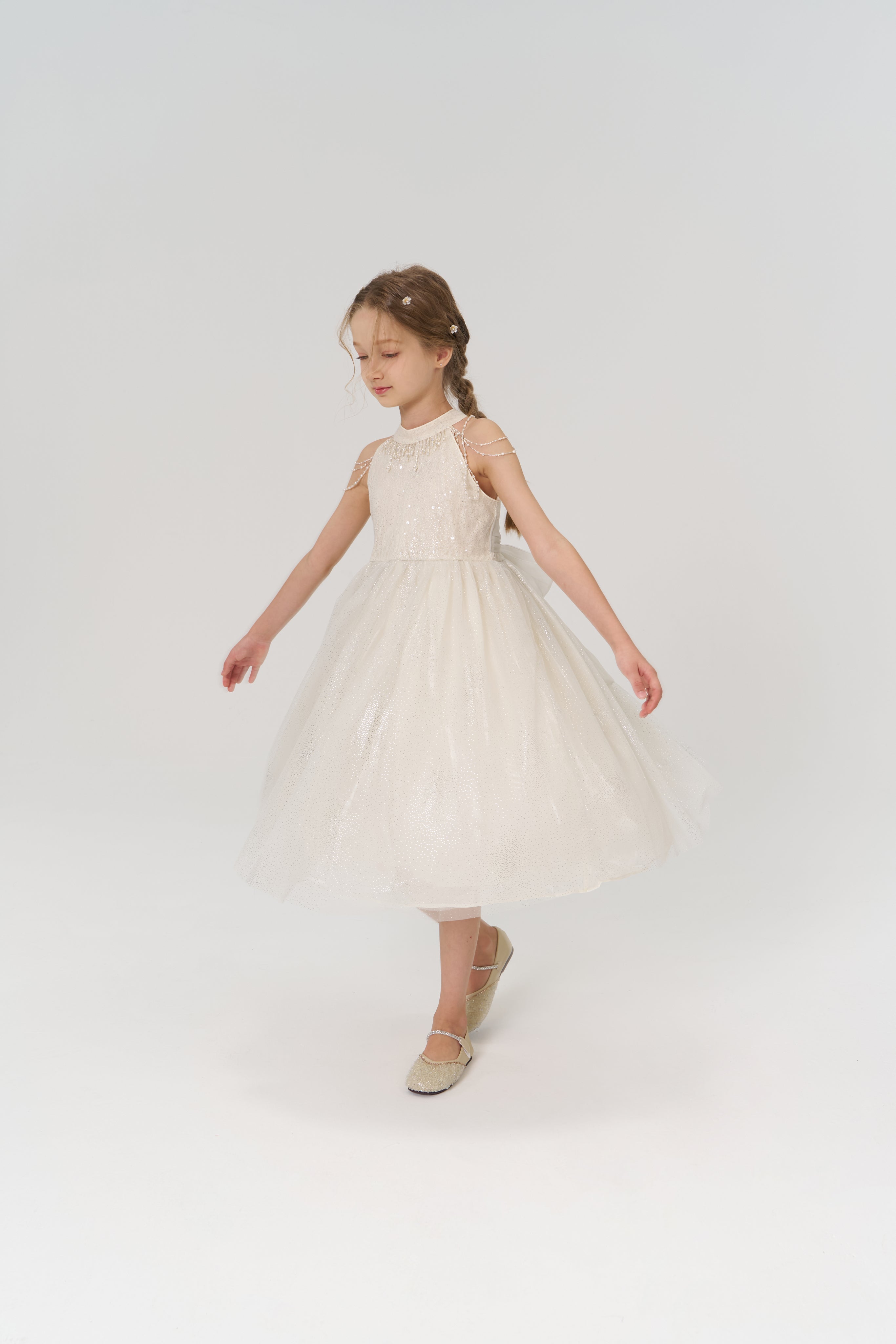 A young girl models the KIKI and JOJO Gilded Whisper Dress—a sleeveless, knee-length beige tulle dress with a delicate skirt—standing against a plain white background.
