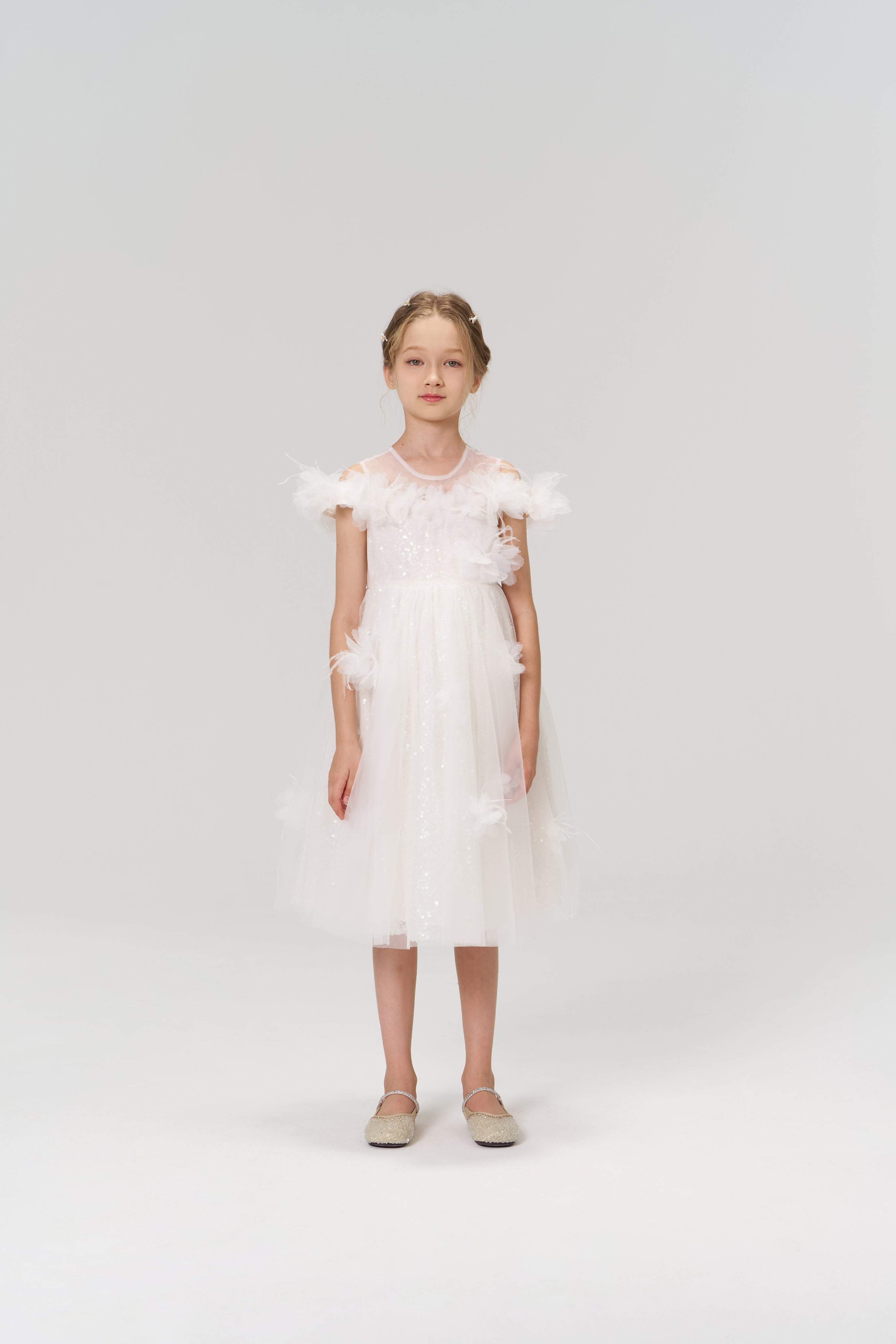 A young girl stands against a plain background wearing the Feathered Whisper Dress by KIKI and JOJO, featuring white sequins, floral feather accents, tulle details, and light-colored shoes.