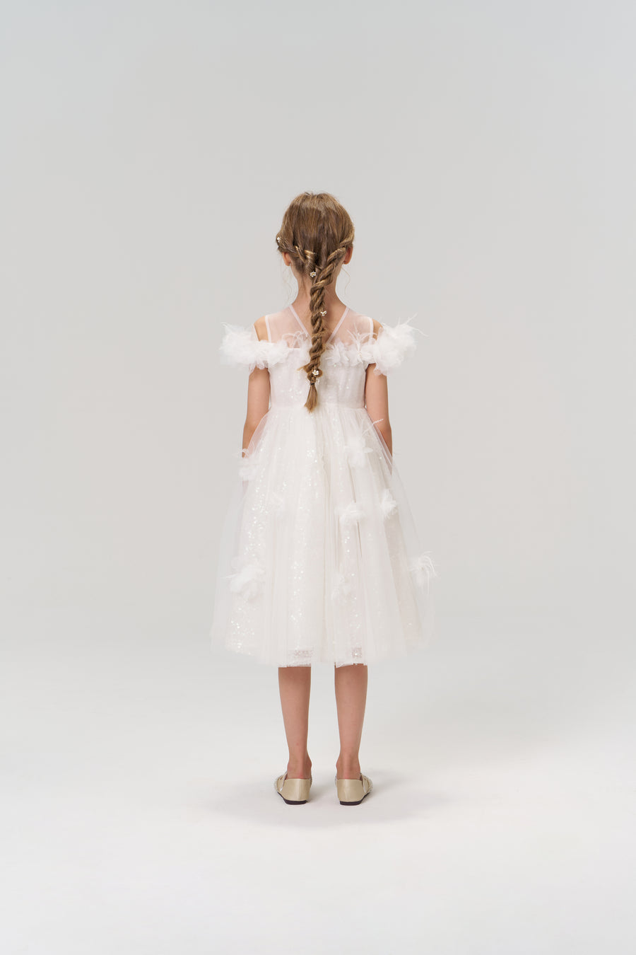 A young girl with braided hair stands facing away, wearing the KIKI and JOJO Feathered Whisper Dress—white with sequin floral details, feather accents, and tulle—paired with gold shoes against a plain light background.
