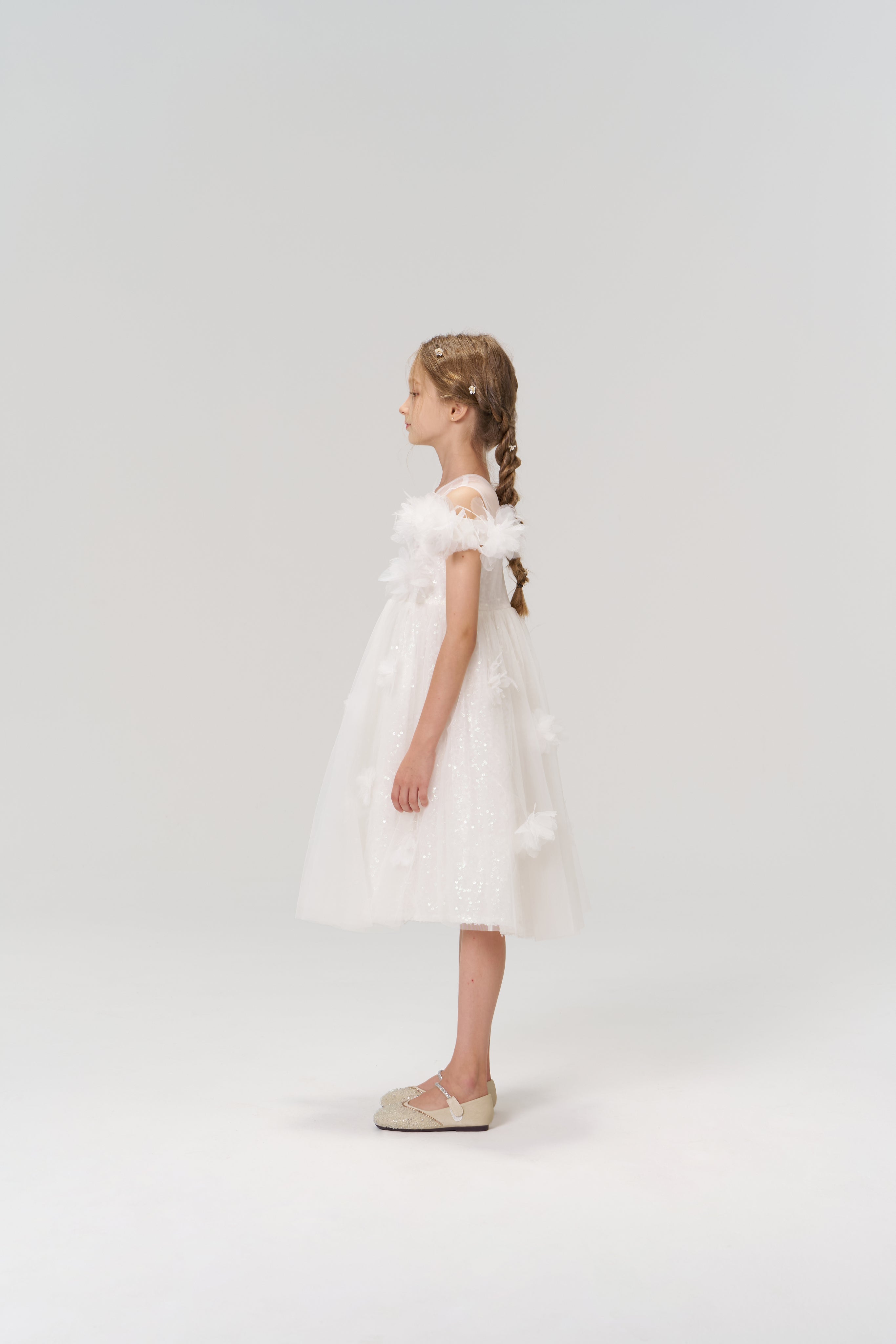 A young girl with braided hair stands in profile against a plain background, wearing the Feathered Whisper Dress by KIKI and JOJO and cream-colored shoes.