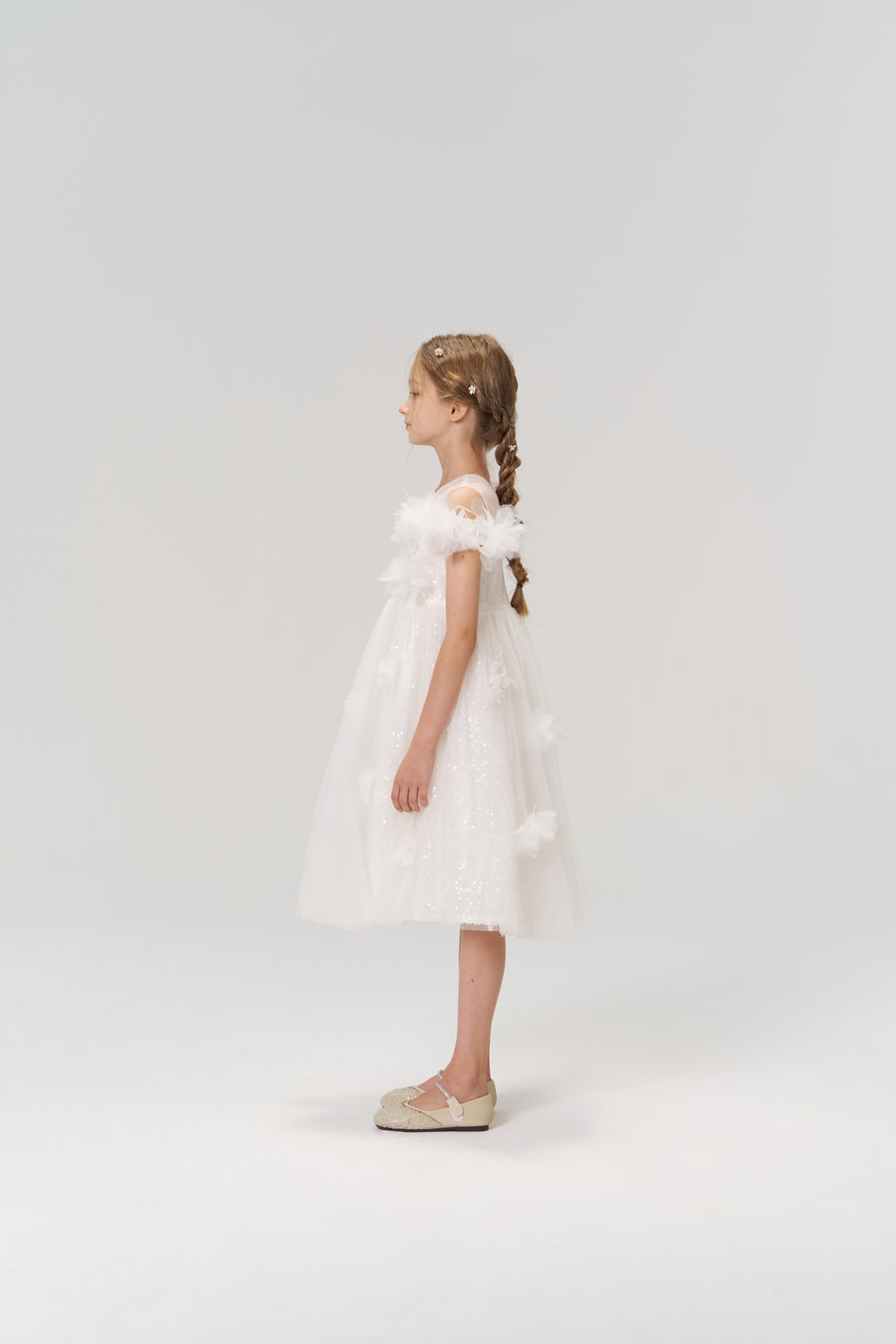 A young girl with braided hair stands in profile against a plain background, wearing the Feathered Whisper Dress by KIKI and JOJO and cream-colored shoes.