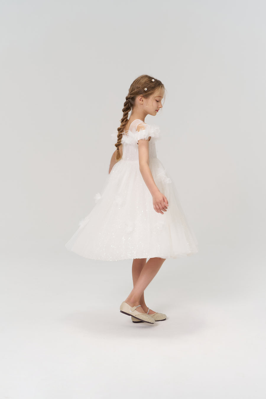 A young girl with braided hair radiates elegance in profile, wearing the KIKI and JOJO Feathered Whisper Dress in blue against a simple light background, paired with light shoes.