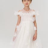 A young girl faces forward in the KIKI and JOJO Feathered Whisper Dress - Blue, a sequin floral feather knee-length tulle piece with delicate embellishments, set against a simple light background.