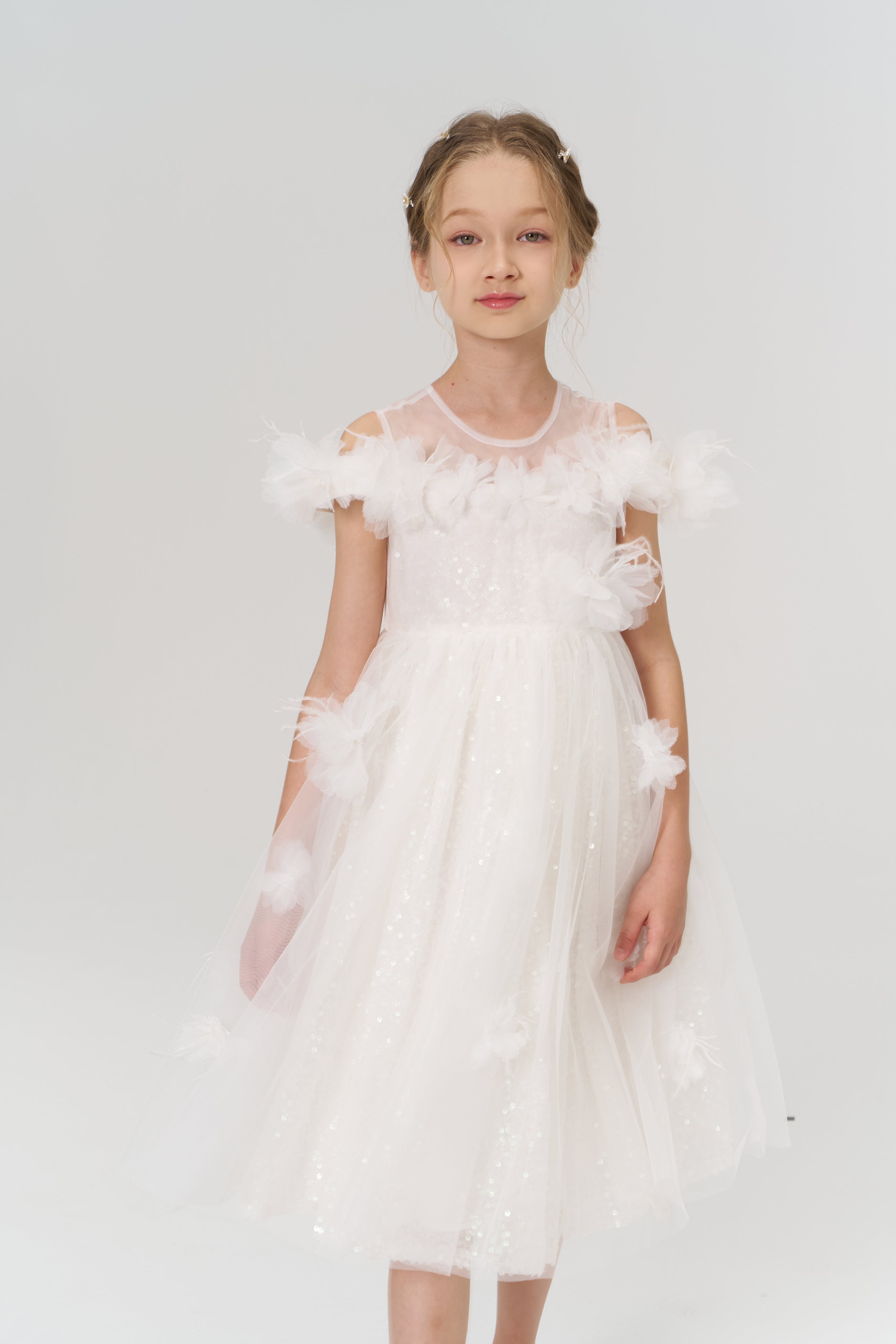 A young girl faces forward in the KIKI and JOJO Feathered Whisper Dress - Blue, a sequin floral feather knee-length tulle piece with delicate embellishments, set against a simple light background.