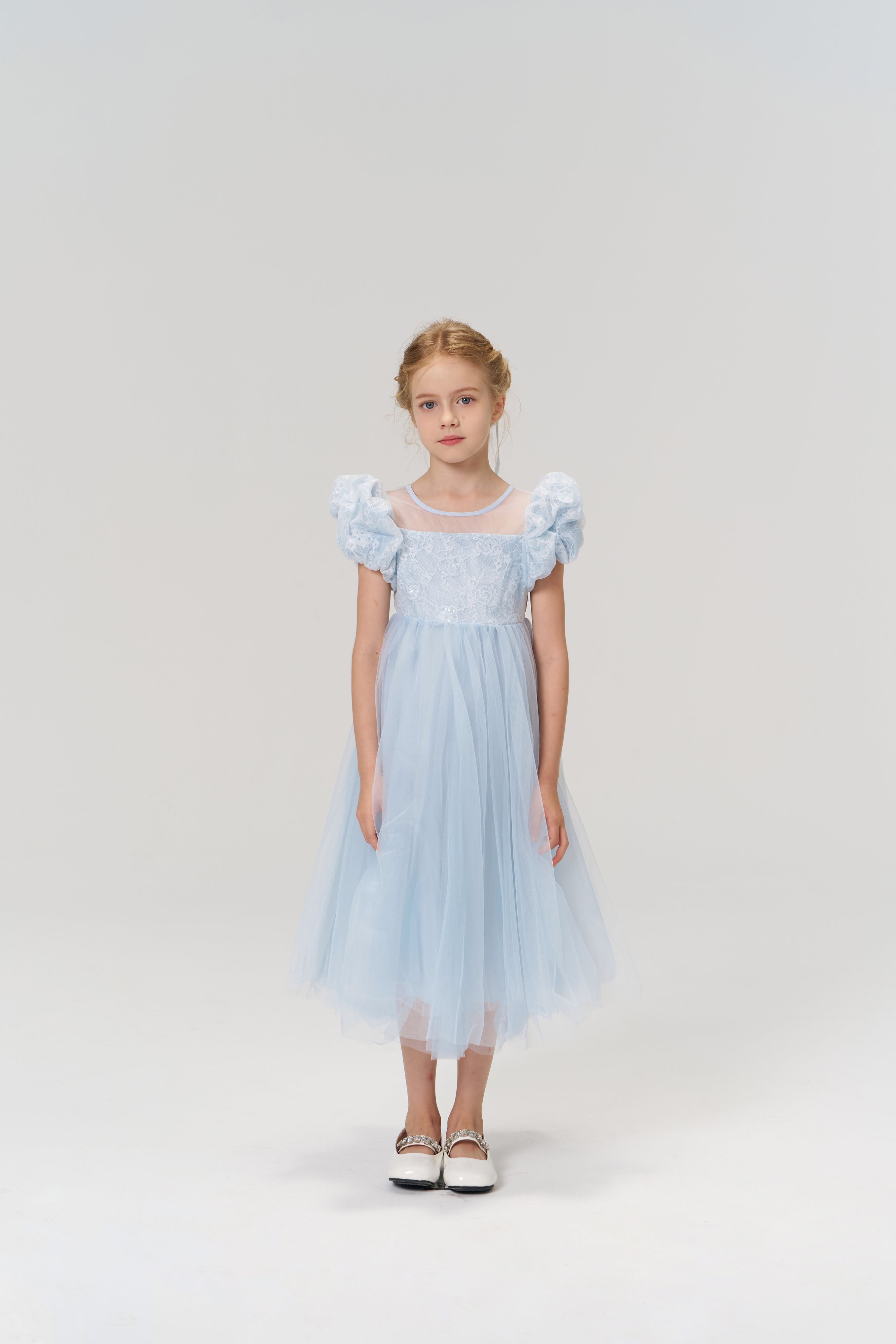 A young girl stands facing forward against a plain, light background, wearing the KIKI and JOJO Sky Whisper Dress—a light blue dress with a tulle skirt—paired with white shoes.