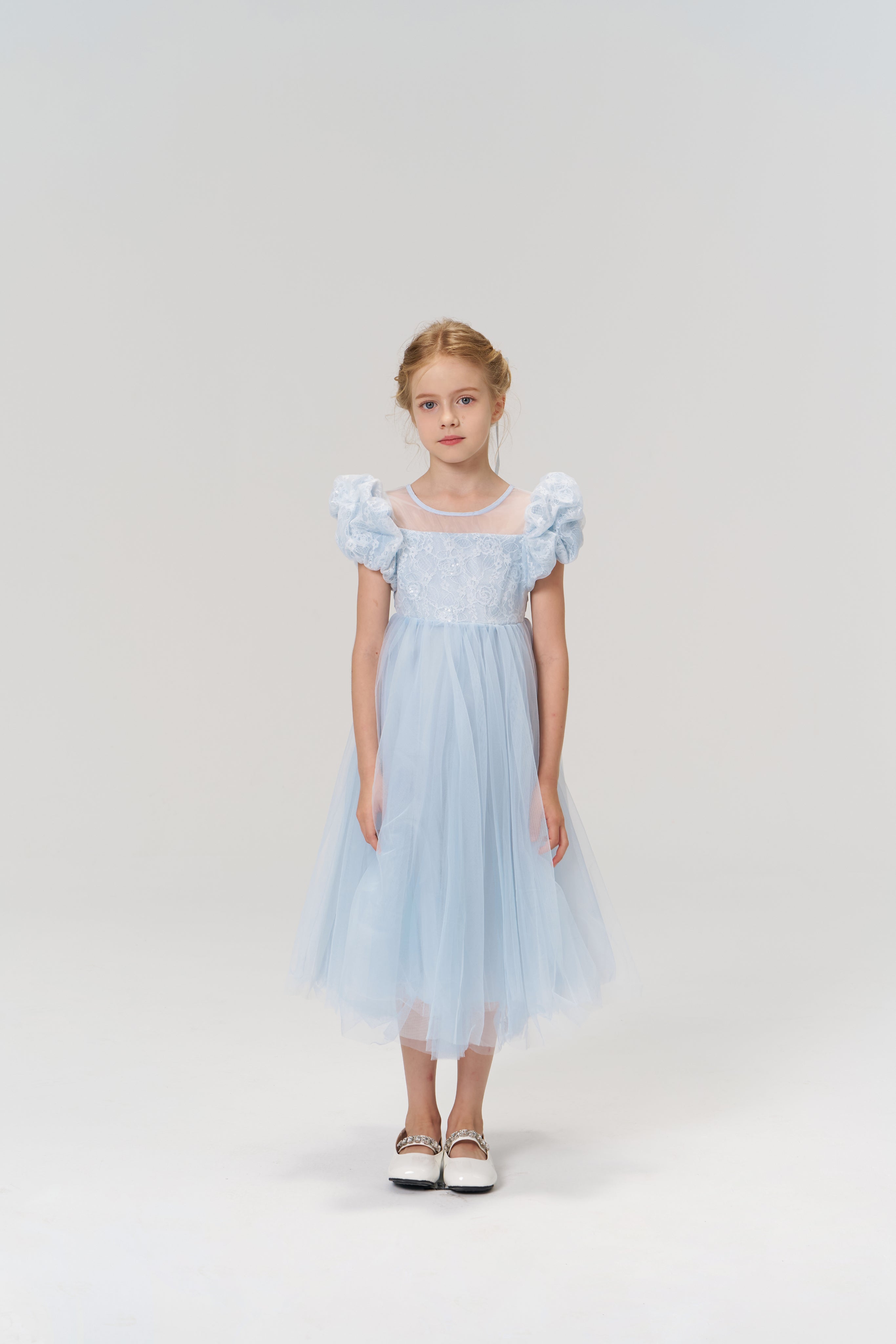 A young girl stands facing forward against a plain, light background, wearing the KIKI and JOJO Sky Whisper Dress—a light blue dress with a tulle skirt—paired with white shoes.