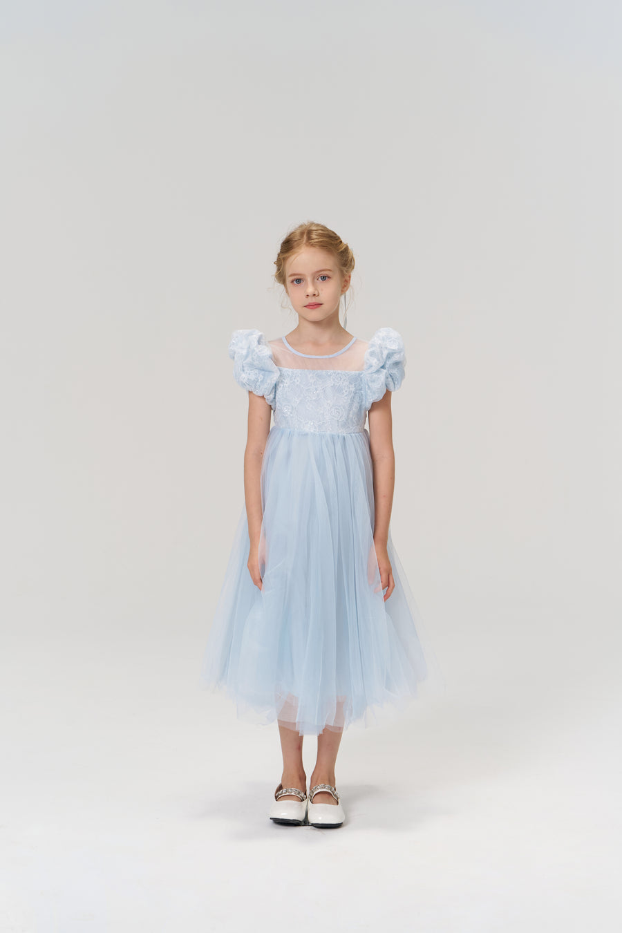A young girl stands facing forward against a plain, light background, wearing the KIKI and JOJO Sky Whisper Dress—a light blue dress with a tulle skirt—paired with white shoes.