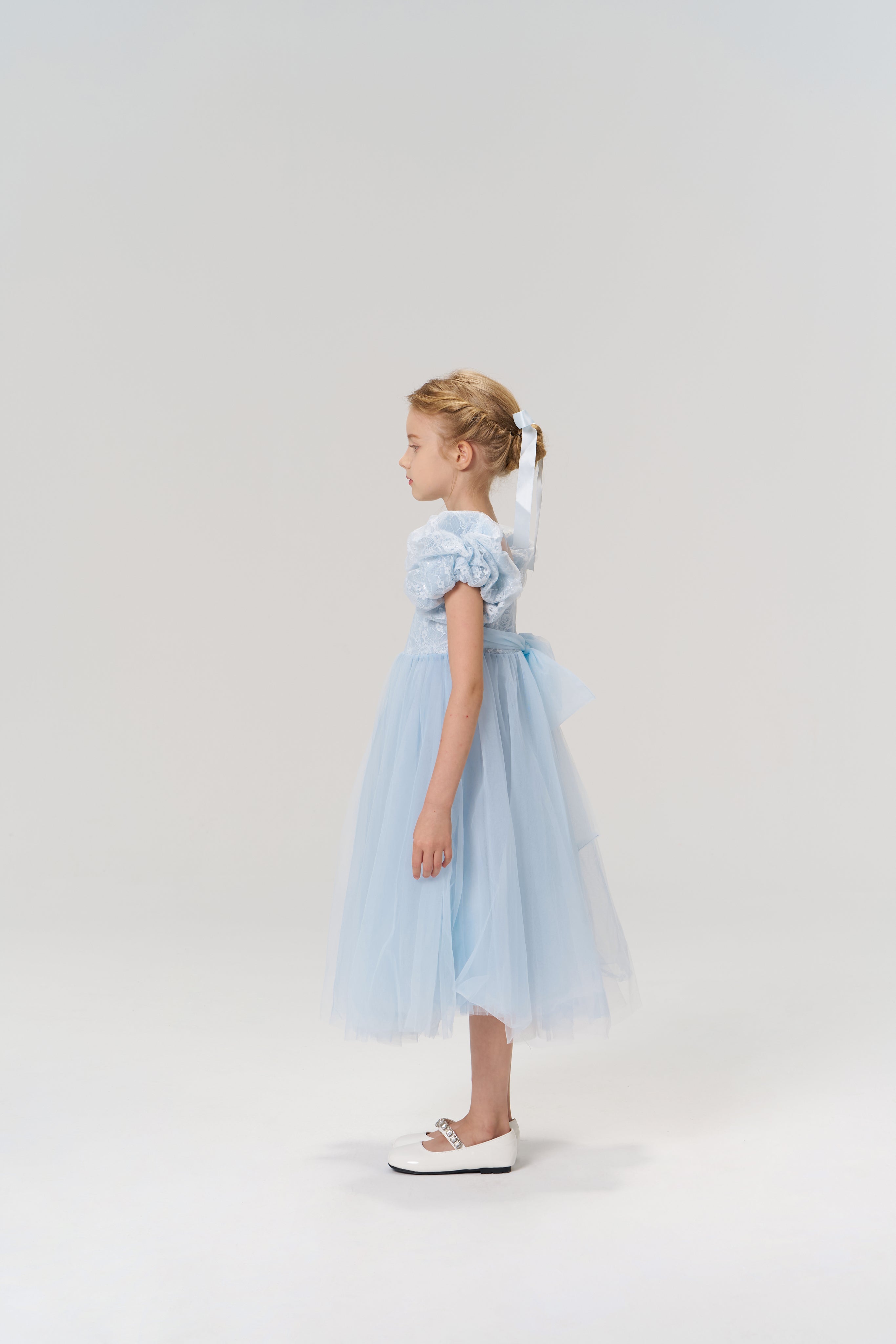 A young girl stands in profile against a plain, light background wearing the Sky Whisper Dress by KIKI and JOJO—a knee-length, light blue lace and organza dress with puffed sleeves—paired with white shoes.