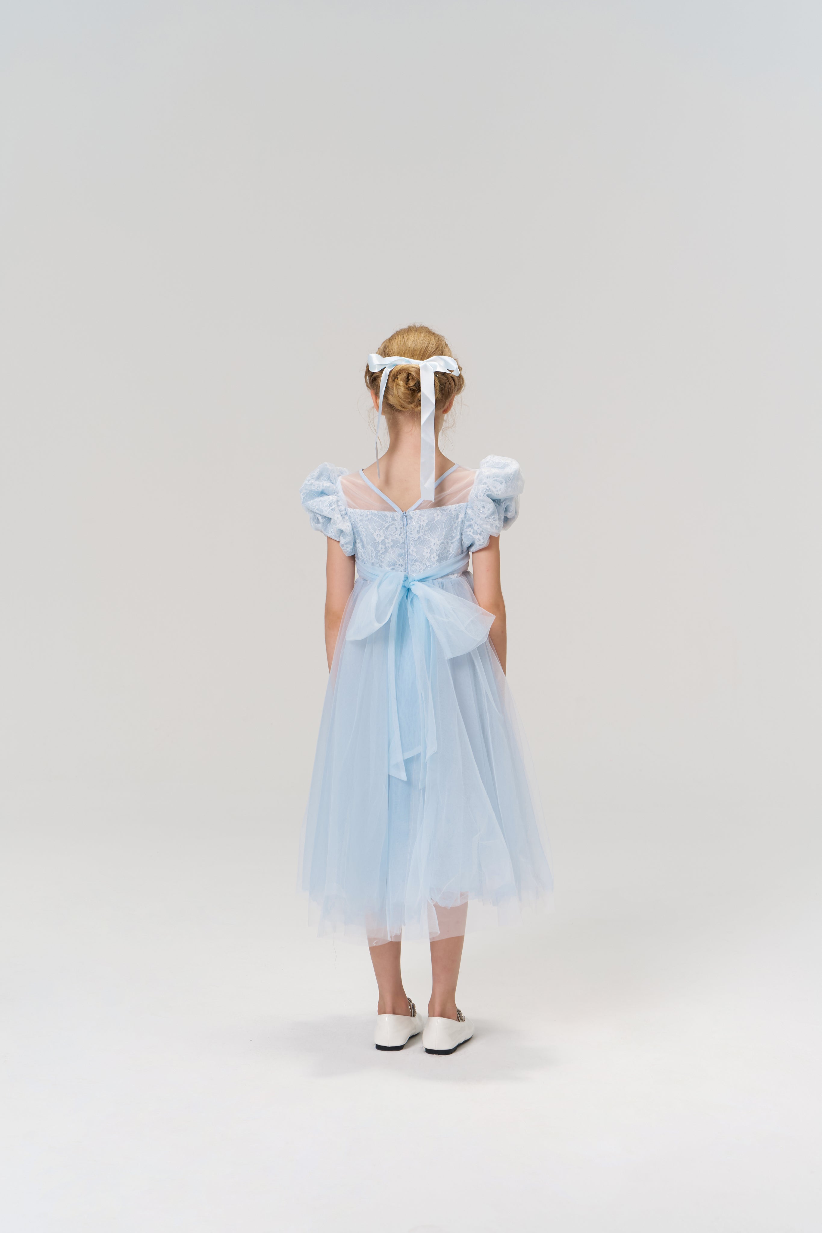 A young girl faces away, wearing the KIKI and JOJO Sky Whisper Dress—light blue lace and organza with puff sleeves, a back bow—paired with white shoes and a ribbon headband, set against a plain background.