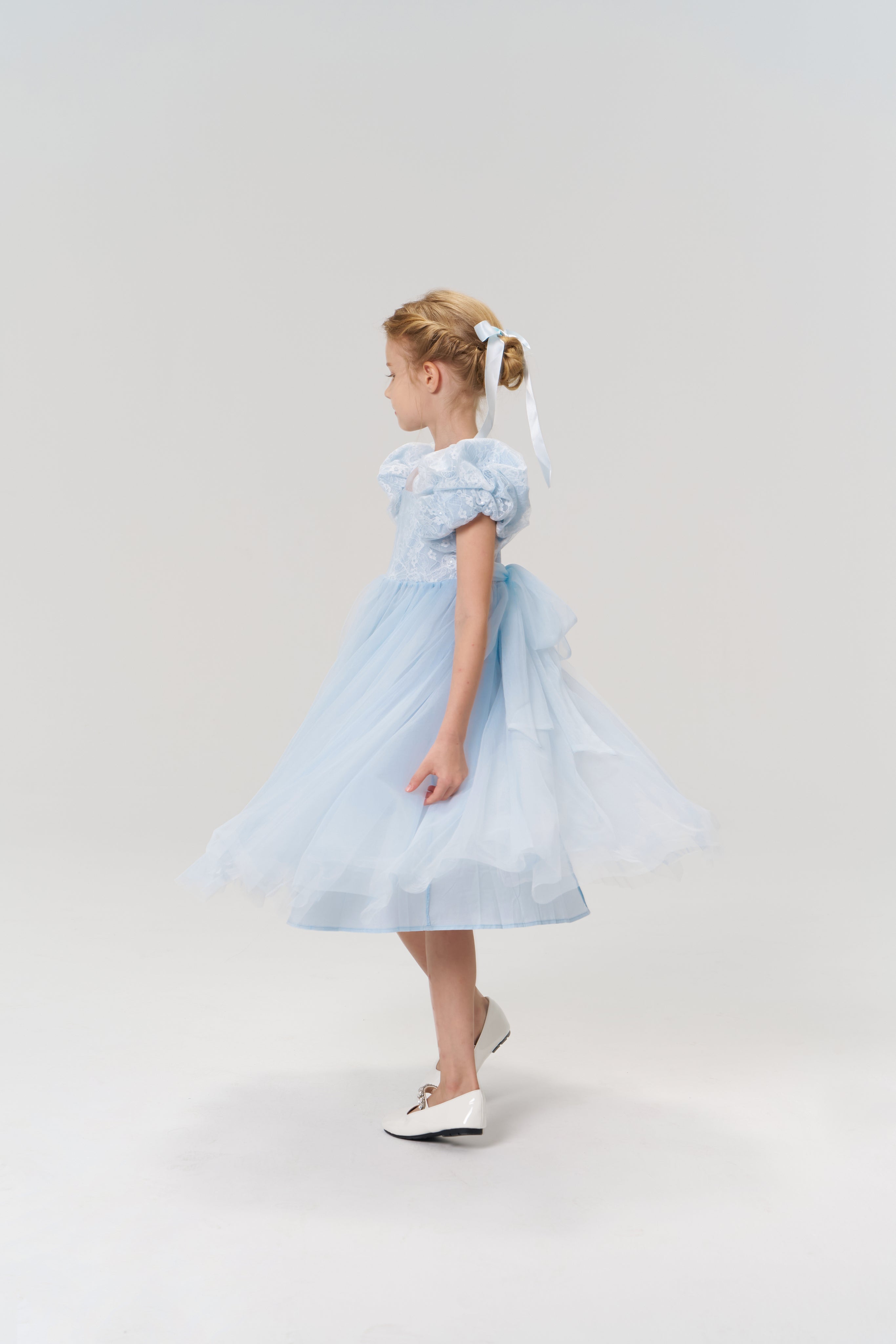 A young girl wearing the Sky Whisper Dress from KIKI and JOJO—light blue with a tulle skirt, puff sleeves, and styled with white ballet flats and a white ribbon in her hair—stands sideways against a plain background.