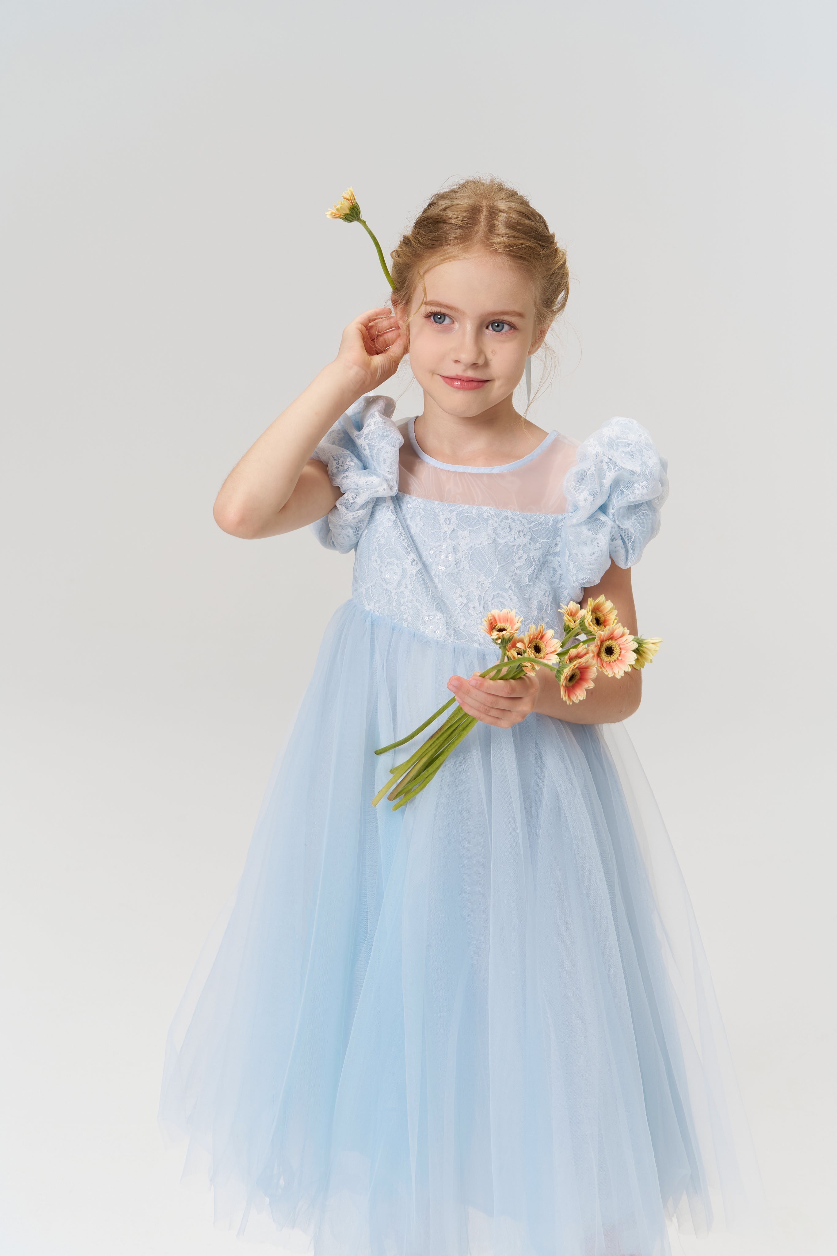 A young girl wears the Sky Whisper Dress by KIKI and JOJO, holding a bouquet and tucking a flower behind her ear, against a plain white background.