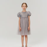 A young girl faces forward in the KIKI and JOJO Dusk Whisper Dress, featuring grey puff sleeves and a glittering tulle skirt, paired with gray shoes against a plain light background.