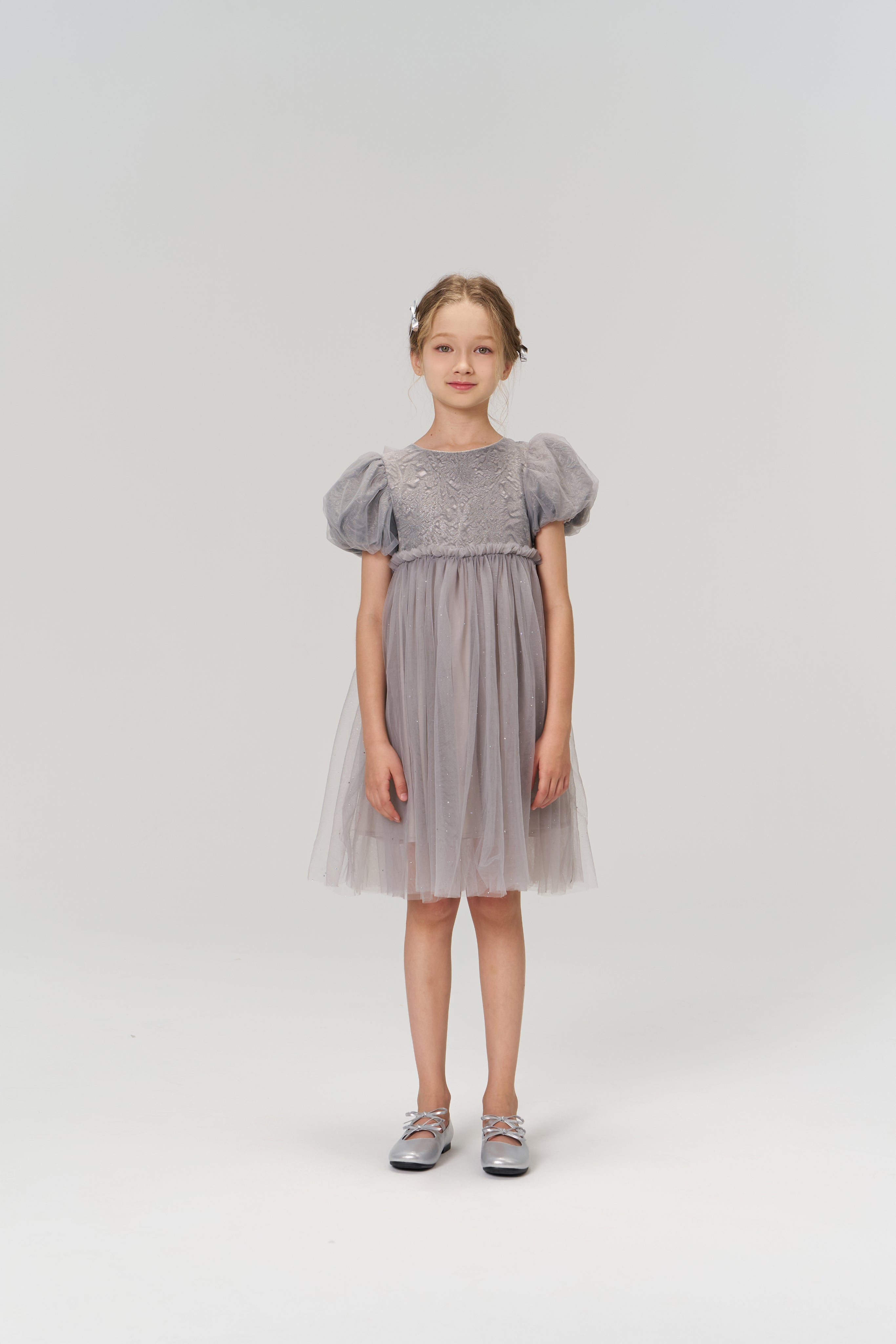 A young girl faces forward in the KIKI and JOJO Dusk Whisper Dress, featuring grey puff sleeves and a glittering tulle skirt, paired with gray shoes against a plain light background.