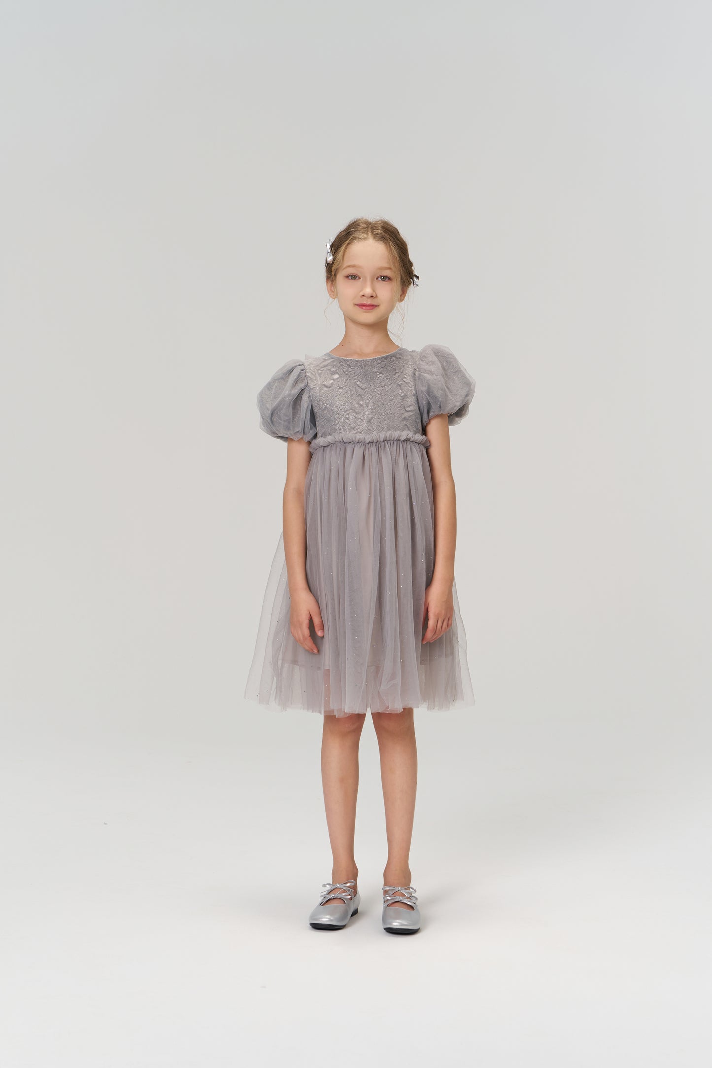 A young girl faces forward in the KIKI and JOJO Dusk Whisper Dress, featuring grey puff sleeves and a glittering tulle skirt, paired with gray shoes against a plain light background.