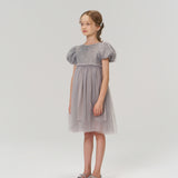A young girl stands in the KIKI and JOJO Dusk Whisper Dress, a grey puff-sleeve style, with silver shoes, facing slightly to the side against a plain light background.