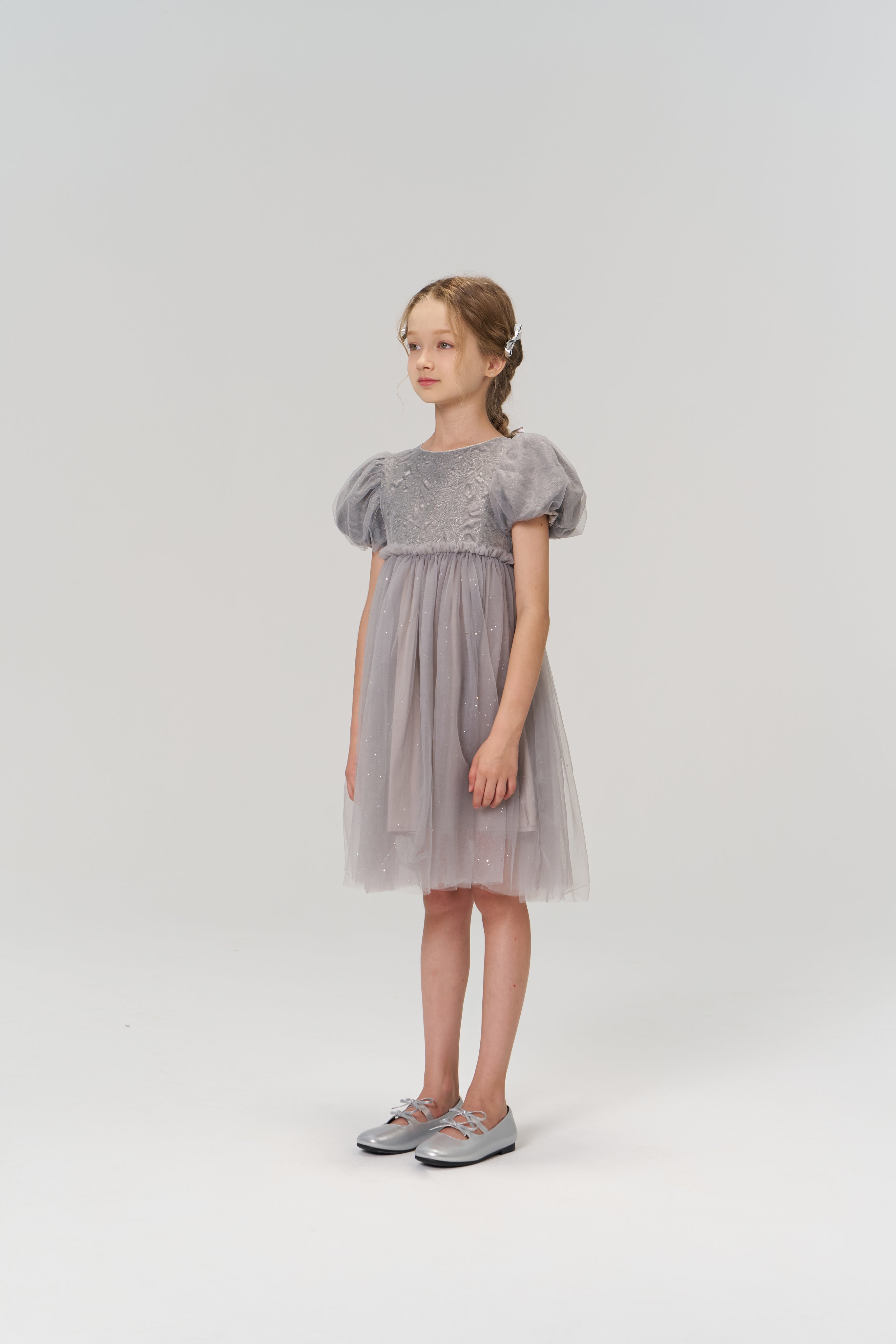 A young girl stands in the KIKI and JOJO Dusk Whisper Dress, a grey puff-sleeve style, with silver shoes, facing slightly to the side against a plain light background.