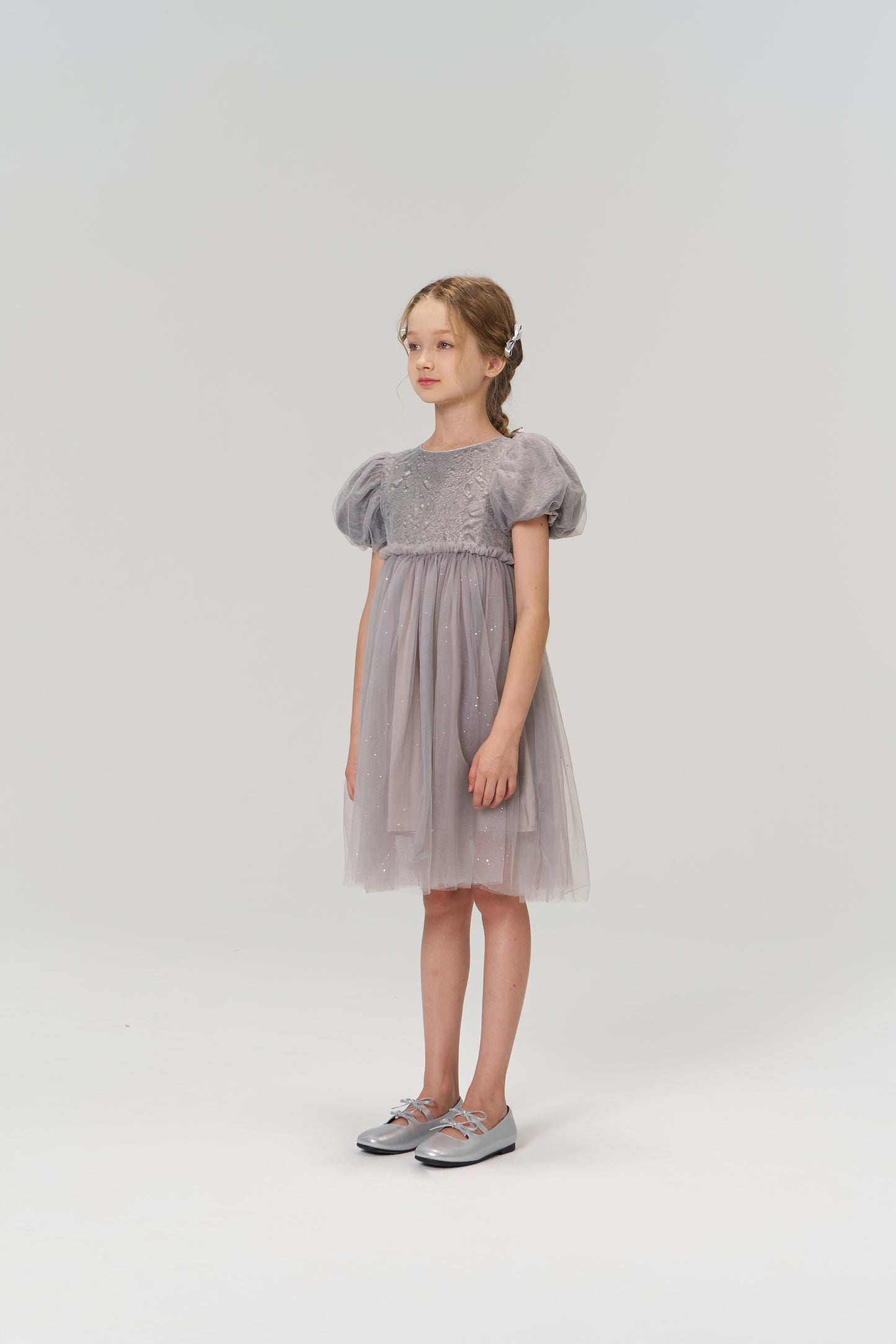 A young girl stands in the KIKI and JOJO Dusk Whisper Dress, a grey puff-sleeve style, with silver shoes, facing slightly to the side against a plain light background.