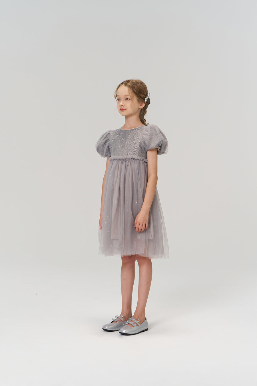 A young girl stands in the KIKI and JOJO Dusk Whisper Dress, a grey puff-sleeve style, with silver shoes, facing slightly to the side against a plain light background.