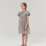 A young girl stands against a plain light background, dressed in the KIKI and JOJO Dusk Whisper Dress with silver shoes, her look radiating sweet elegance and charm.