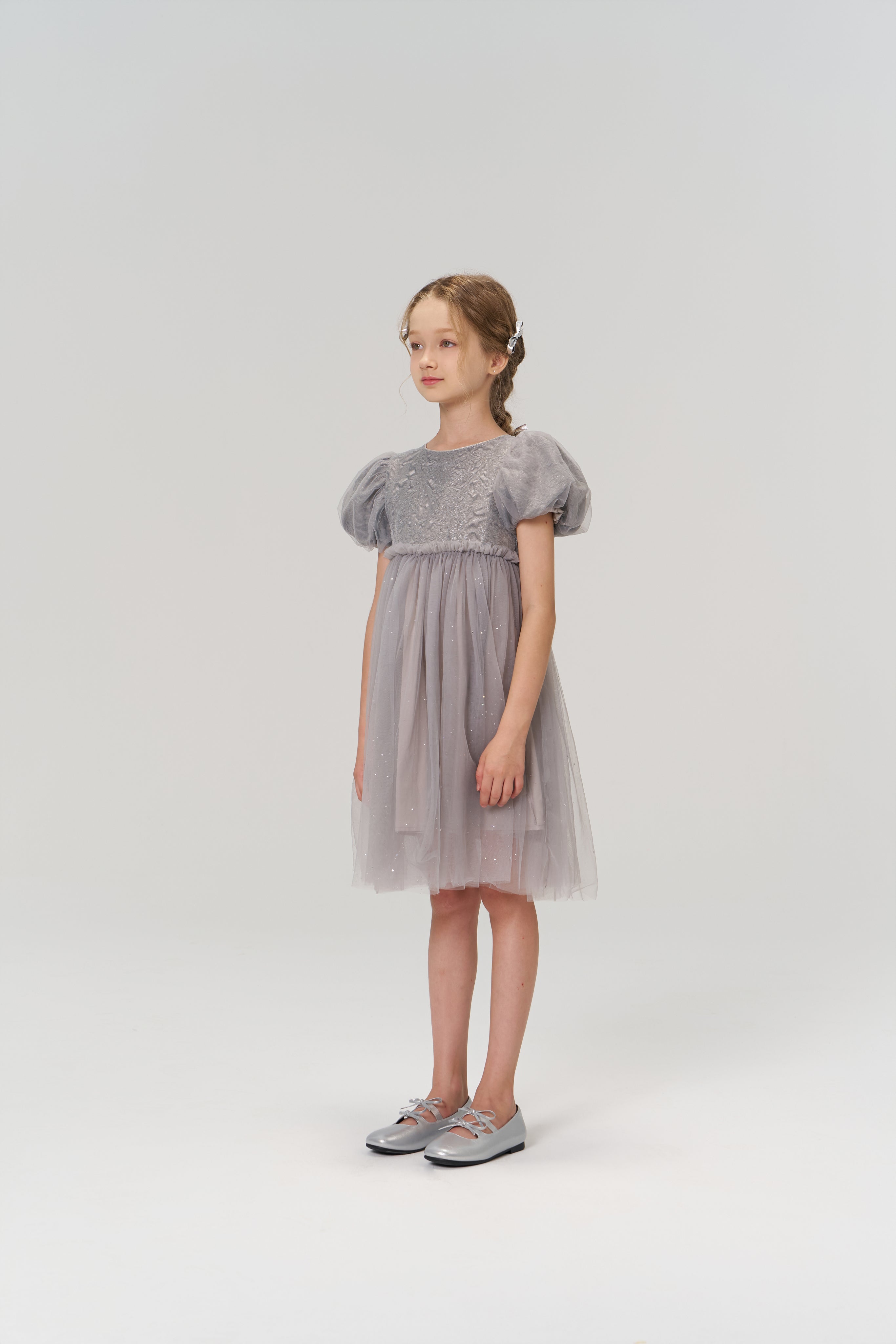 A young girl stands against a plain light background, dressed in the KIKI and JOJO Dusk Whisper Dress with silver shoes, her look radiating sweet elegance and charm.