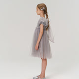 A young girl with braided hair stands in profile, wearing the KIKI and JOJO Dusk Whisper Dress and silver shoes, against a plain white background.