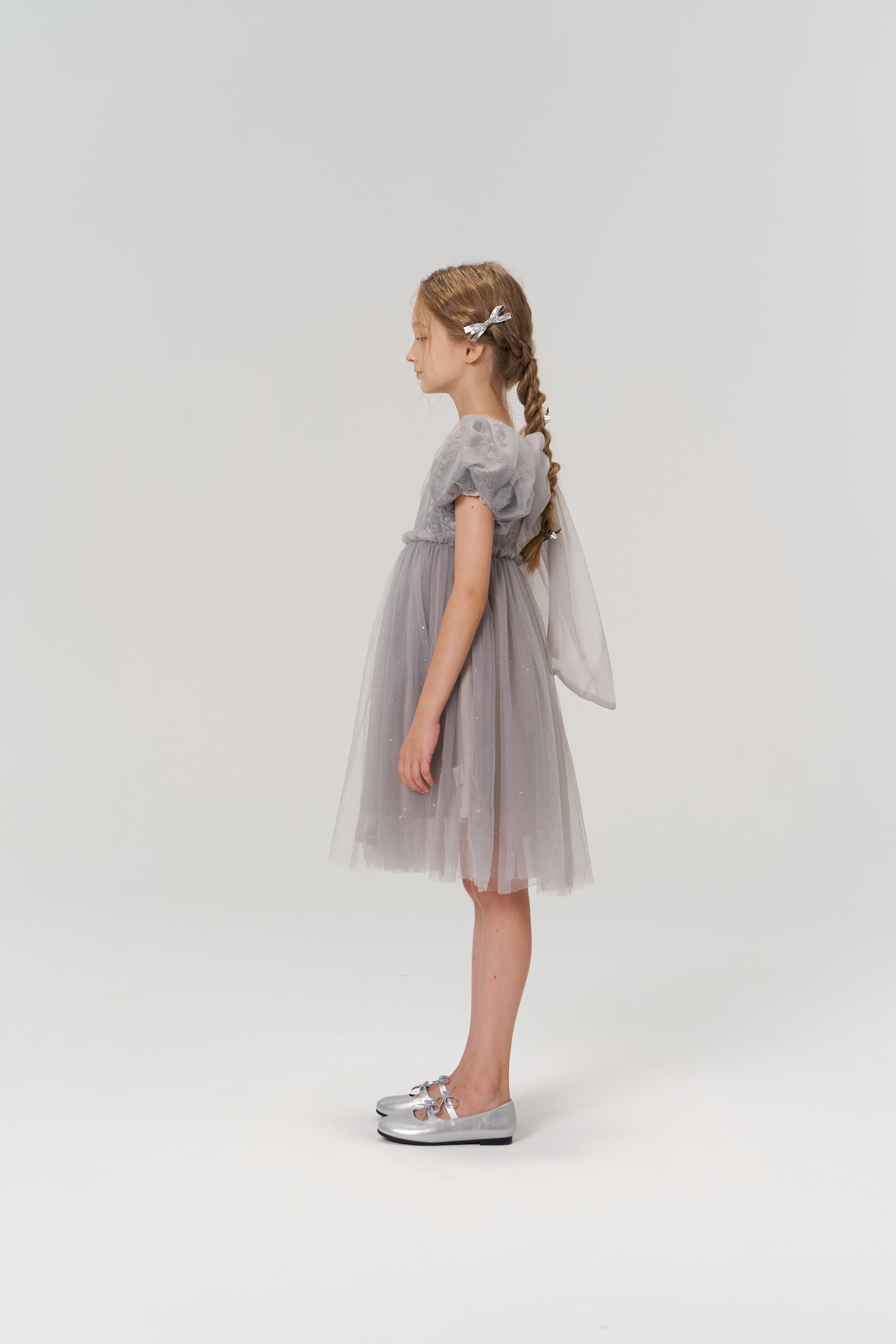A young girl with braided hair stands in profile, wearing the KIKI and JOJO Dusk Whisper Dress and silver shoes, against a plain white background.