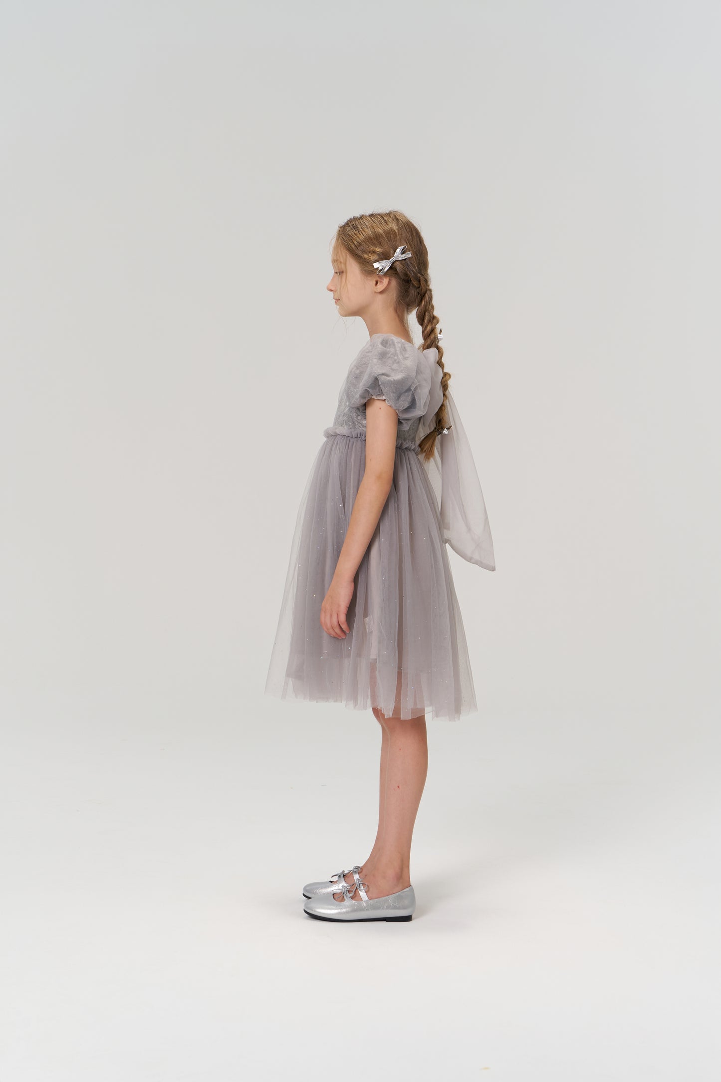A young girl with braided hair stands in profile, wearing the KIKI and JOJO Dusk Whisper Dress and silver shoes, against a plain white background.