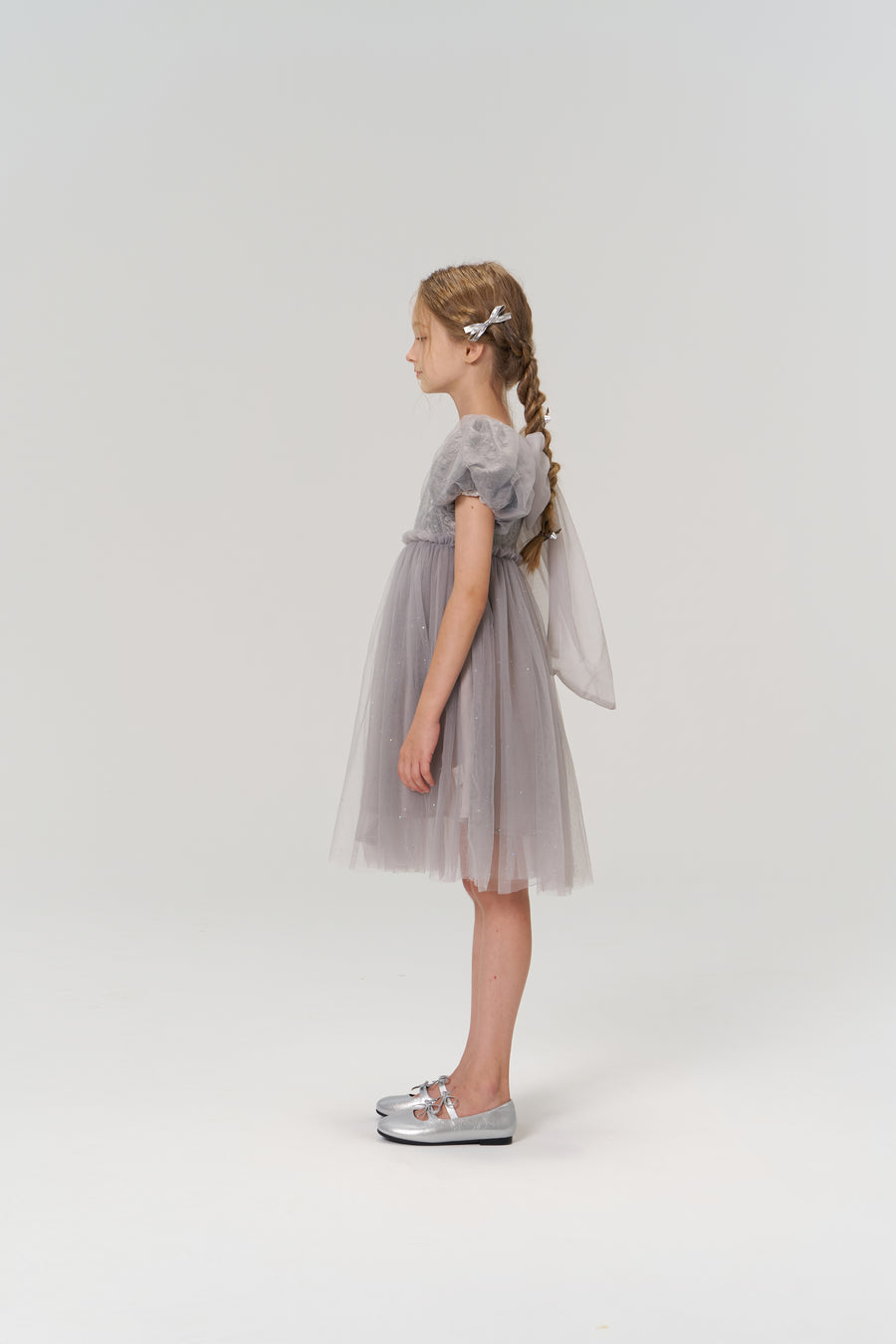 A young girl with braided hair stands in profile, wearing the KIKI and JOJO Dusk Whisper Dress and silver shoes, against a plain white background.