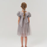 A young girl with braided hair faces away, wearing the KIKI and JOJO Dusk Whisper Dress—a grey puff-sleeve style with sheer fabric—and silver shoes against a plain light background.