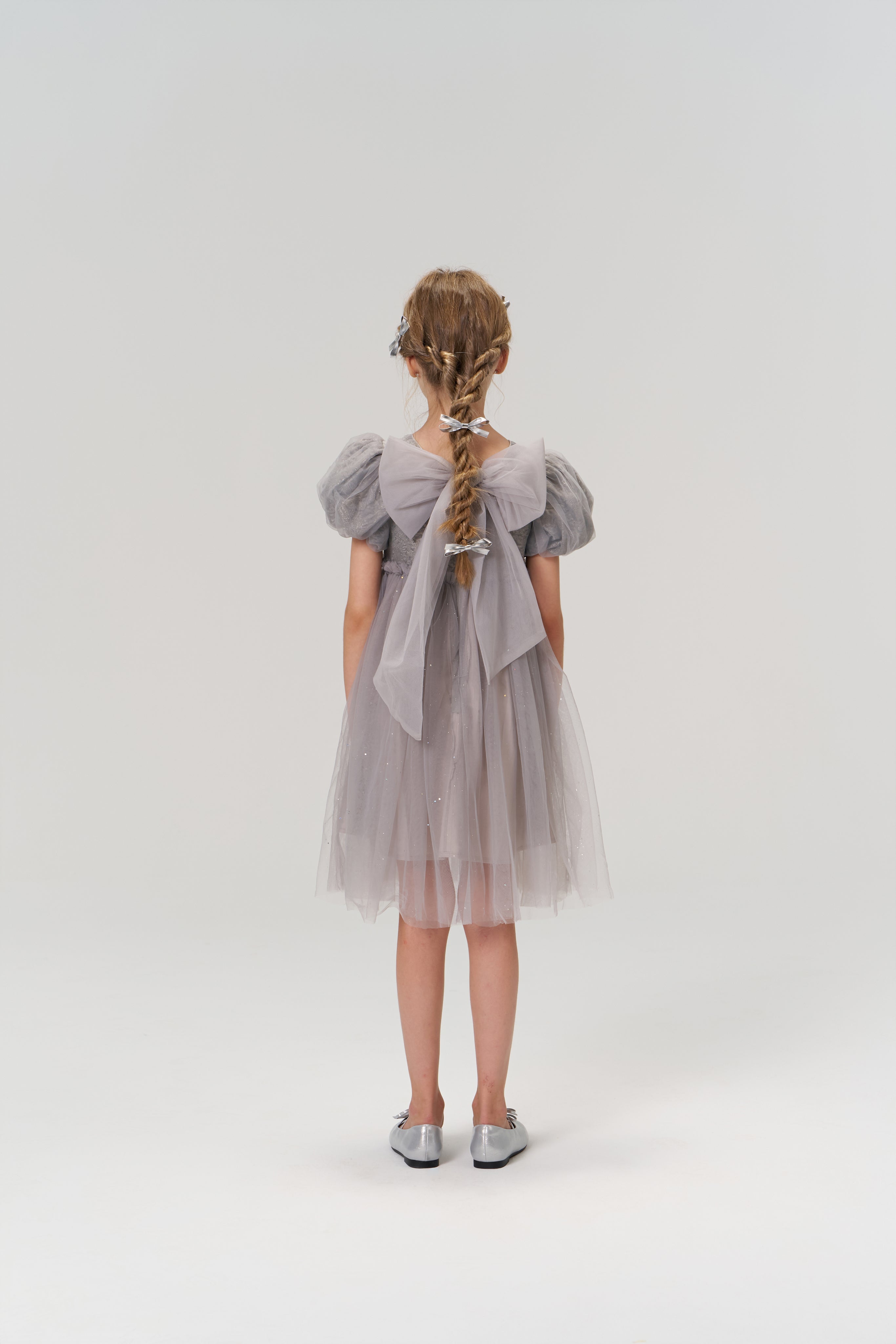 A young girl with braided hair faces away, wearing the KIKI and JOJO Dusk Whisper Dress—a grey puff-sleeve style with sheer fabric—and silver shoes against a plain light background.