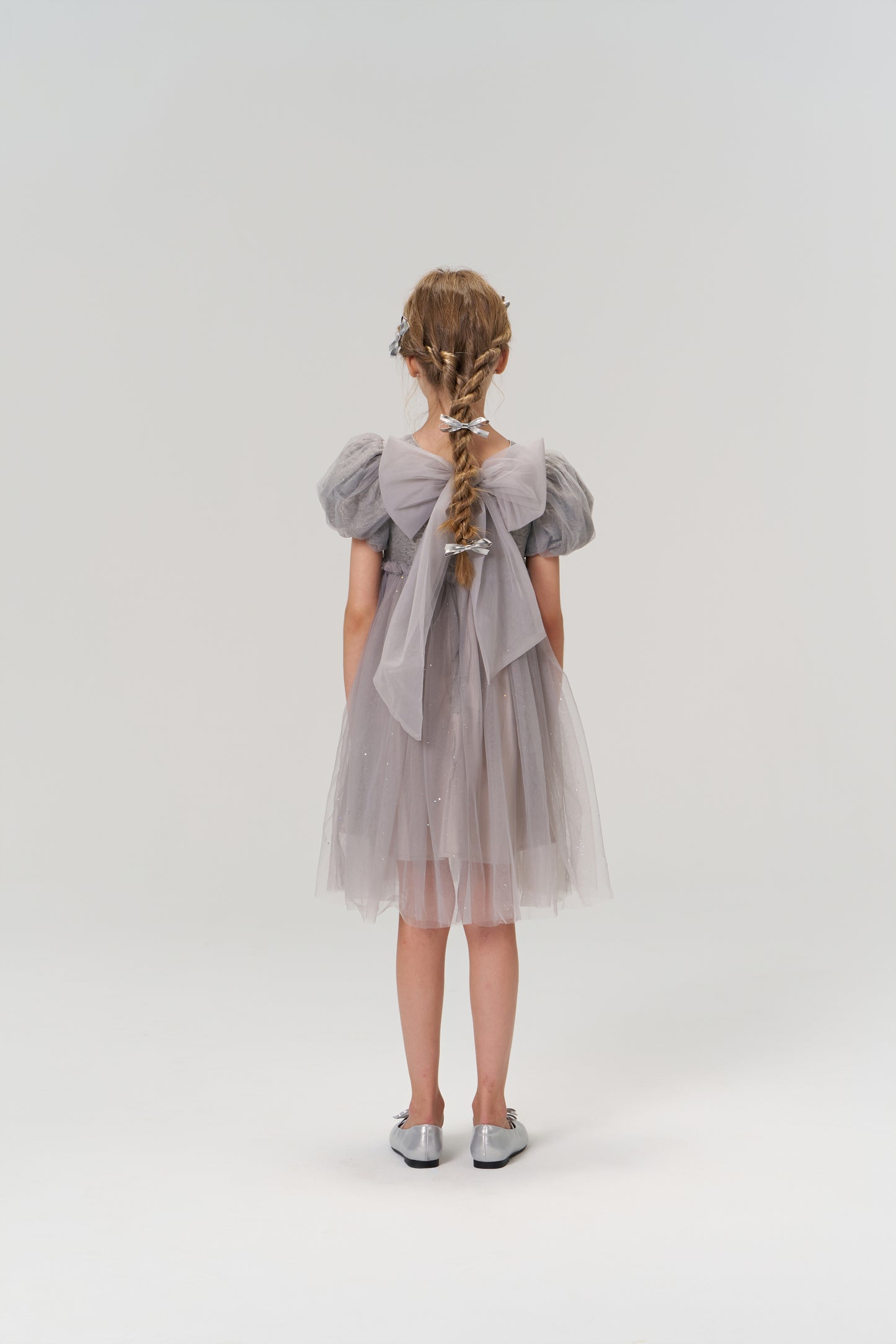 A young girl with braided hair faces away, wearing the KIKI and JOJO Dusk Whisper Dress—a grey puff-sleeve style with sheer fabric—and silver shoes against a plain light background.