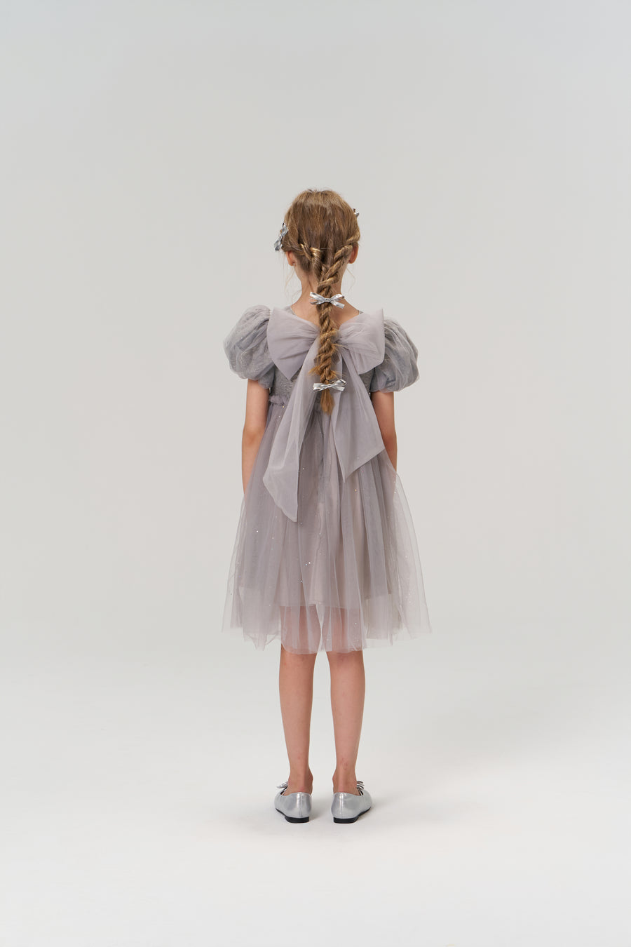 A young girl with braided hair faces away, wearing the KIKI and JOJO Dusk Whisper Dress—a grey puff-sleeve style with sheer fabric—and silver shoes against a plain light background.