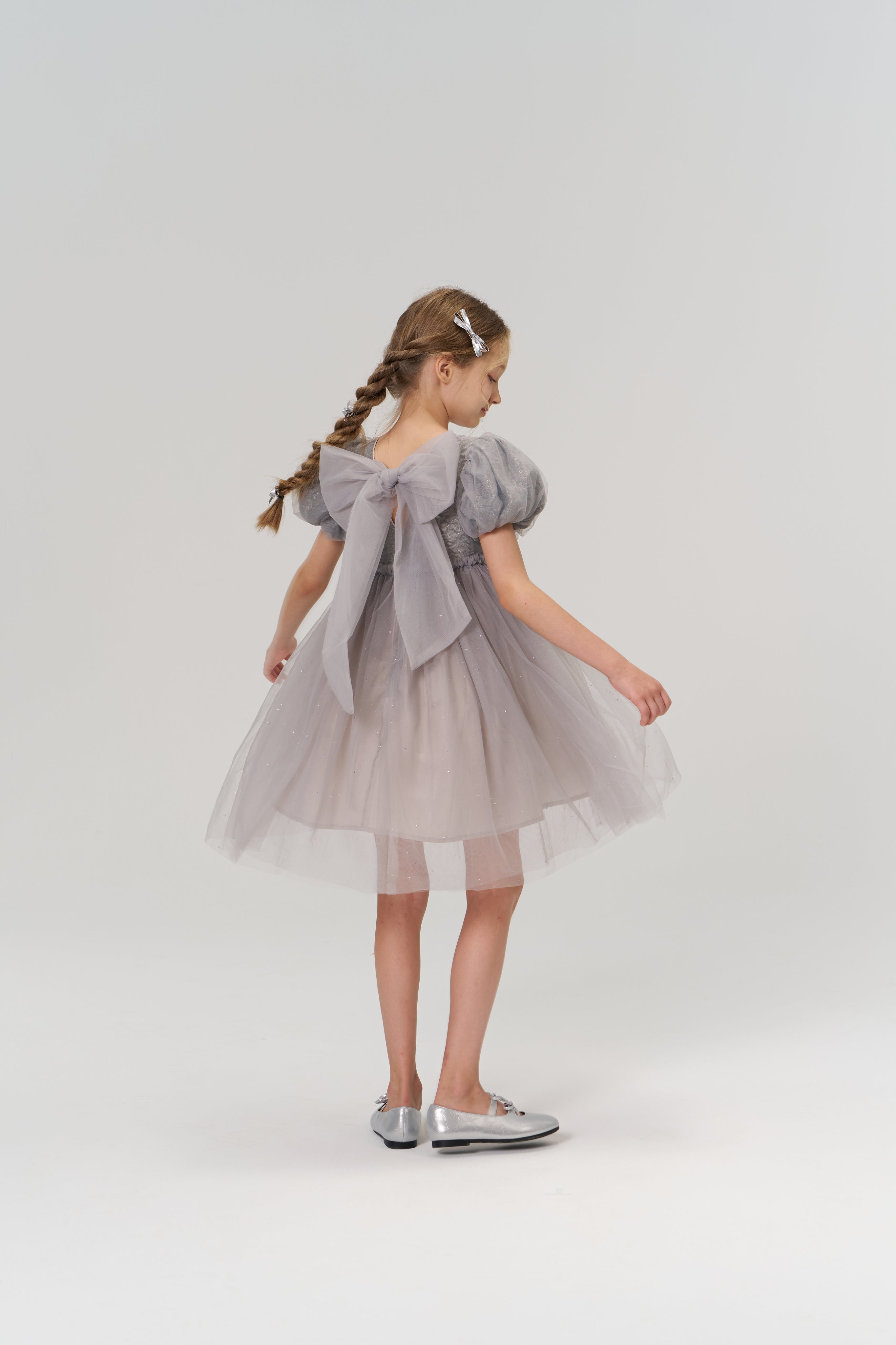A young girl with braided hair styled with KIKI and JOJO Girls Silver Bow Hair Clips (Set of 2) wears a gray tulle dress with a large back bow and silver shoes, standing against a plain light background.