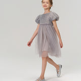 A young girl wearing the Dusk Whisper Dress by KIKI and JOJO and silver shoes walks forward against a plain light background.