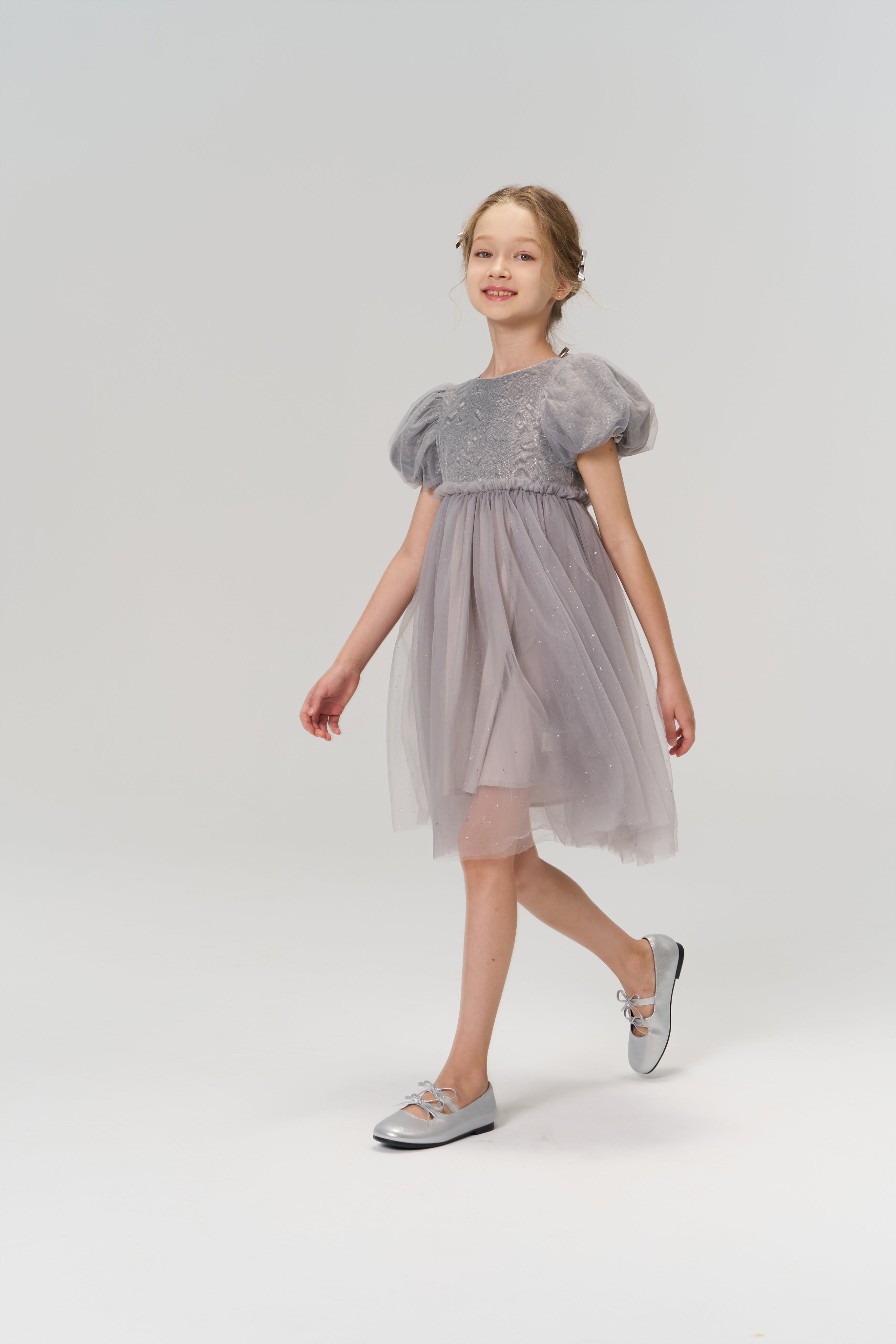 A young girl wearing the Dusk Whisper Dress by KIKI and JOJO and silver shoes walks forward against a plain light background.