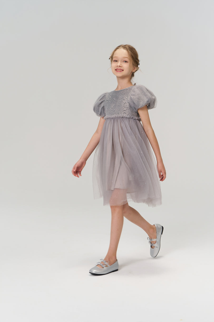 A young girl wearing the Dusk Whisper Dress by KIKI and JOJO and silver shoes walks forward against a plain light background.