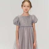 A young girl with light brown hair wears the KIKI and JOJO Dusk Whisper Dress, featuring grey puff sleeves, as she stands against a plain light background and gazes slightly to the side.