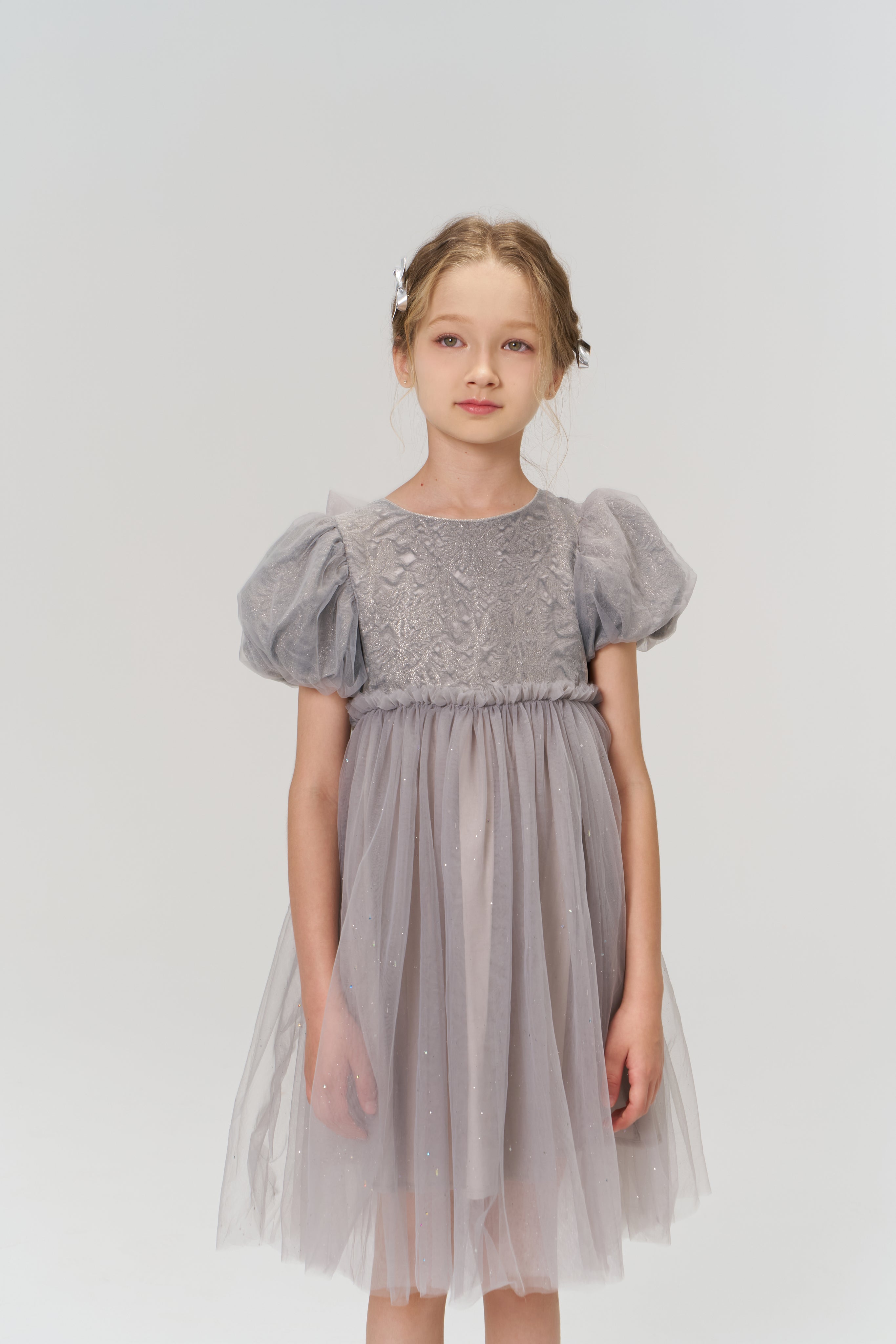 A young girl with light brown hair wears the KIKI and JOJO Dusk Whisper Dress, featuring grey puff sleeves, as she stands against a plain light background and gazes slightly to the side.