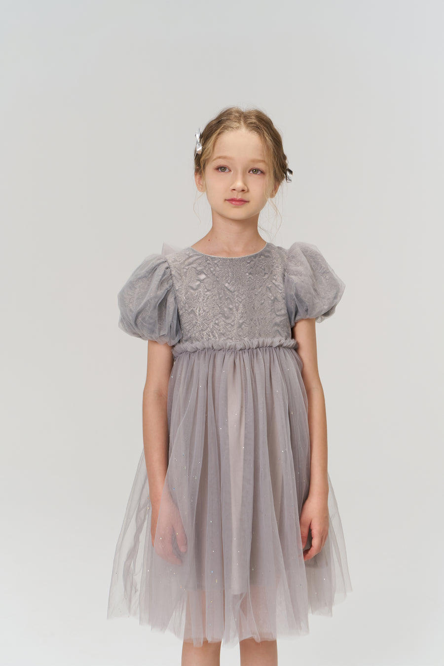A young girl with light brown hair wears the KIKI and JOJO Dusk Whisper Dress, featuring grey puff sleeves, as she stands against a plain light background and gazes slightly to the side.