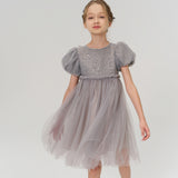 A young girl in the KIKI and JOJO Dusk Whisper Dress—featuring grey puff sleeves and glittering tulle—pairs it with silver shoes, standing against a plain white background.