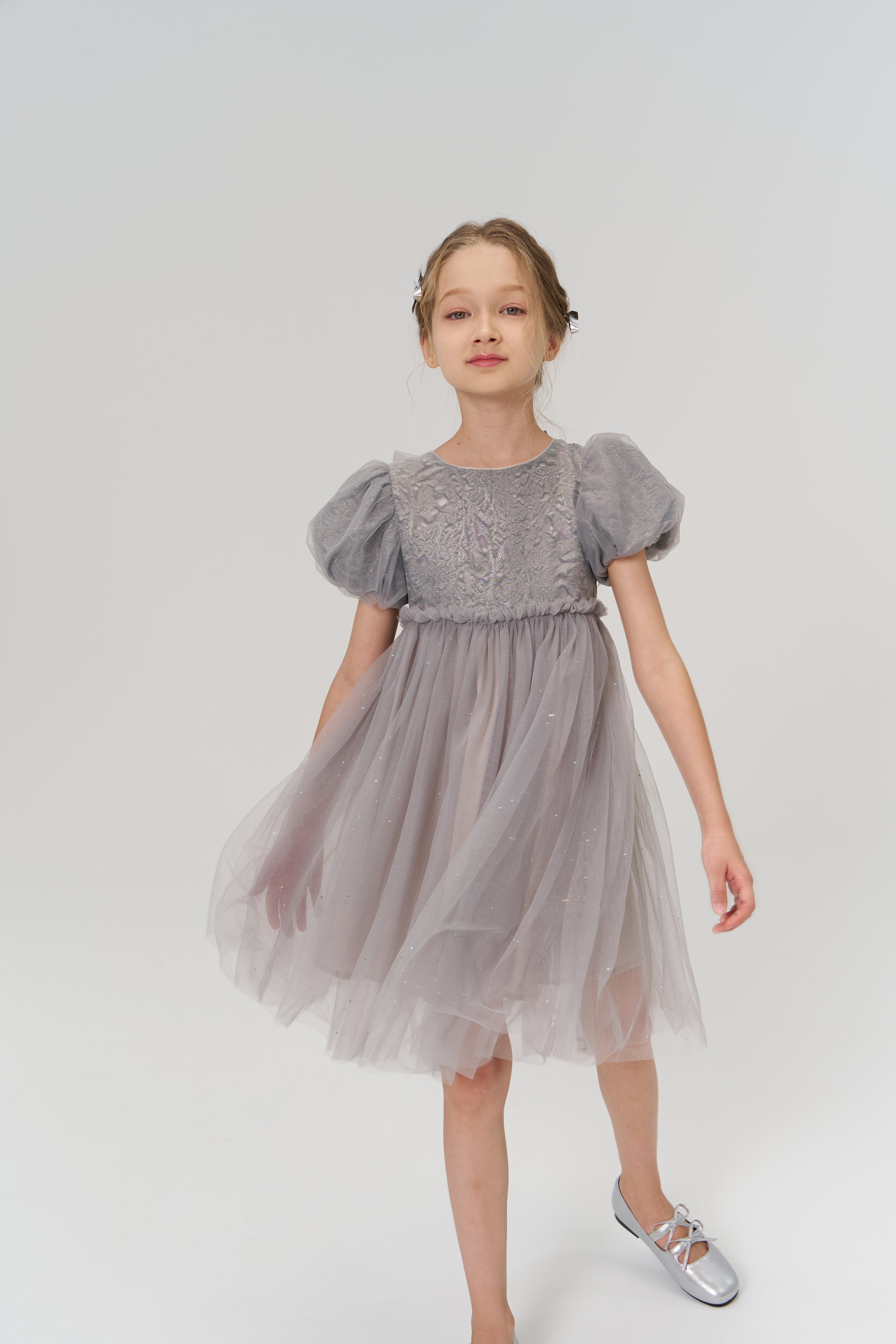 A young girl in the KIKI and JOJO Dusk Whisper Dress—featuring grey puff sleeves and glittering tulle—pairs it with silver shoes, standing against a plain white background.