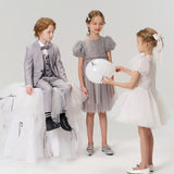 Three children pose against a plain backdrop: a boy in a suit sits on a white tulle platform, while one girl wears the KIKI and JOJO Dusk Whisper Dress and another dons an A-line babydoll dress, both playing with a white balloon.