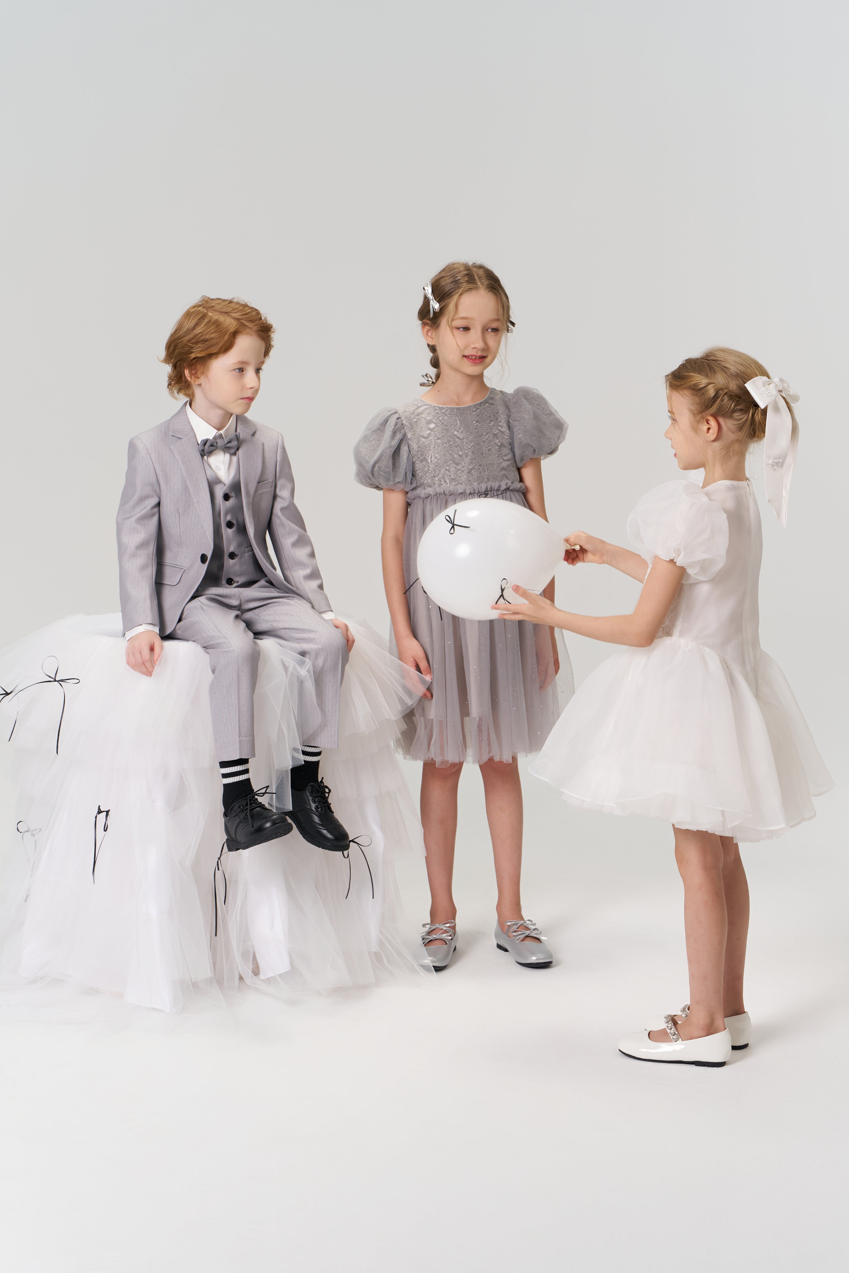 Three children pose against a plain backdrop: a boy in a suit sits on a white tulle platform, while one girl wears the KIKI and JOJO Dusk Whisper Dress and another dons an A-line babydoll dress, both playing with a white balloon.