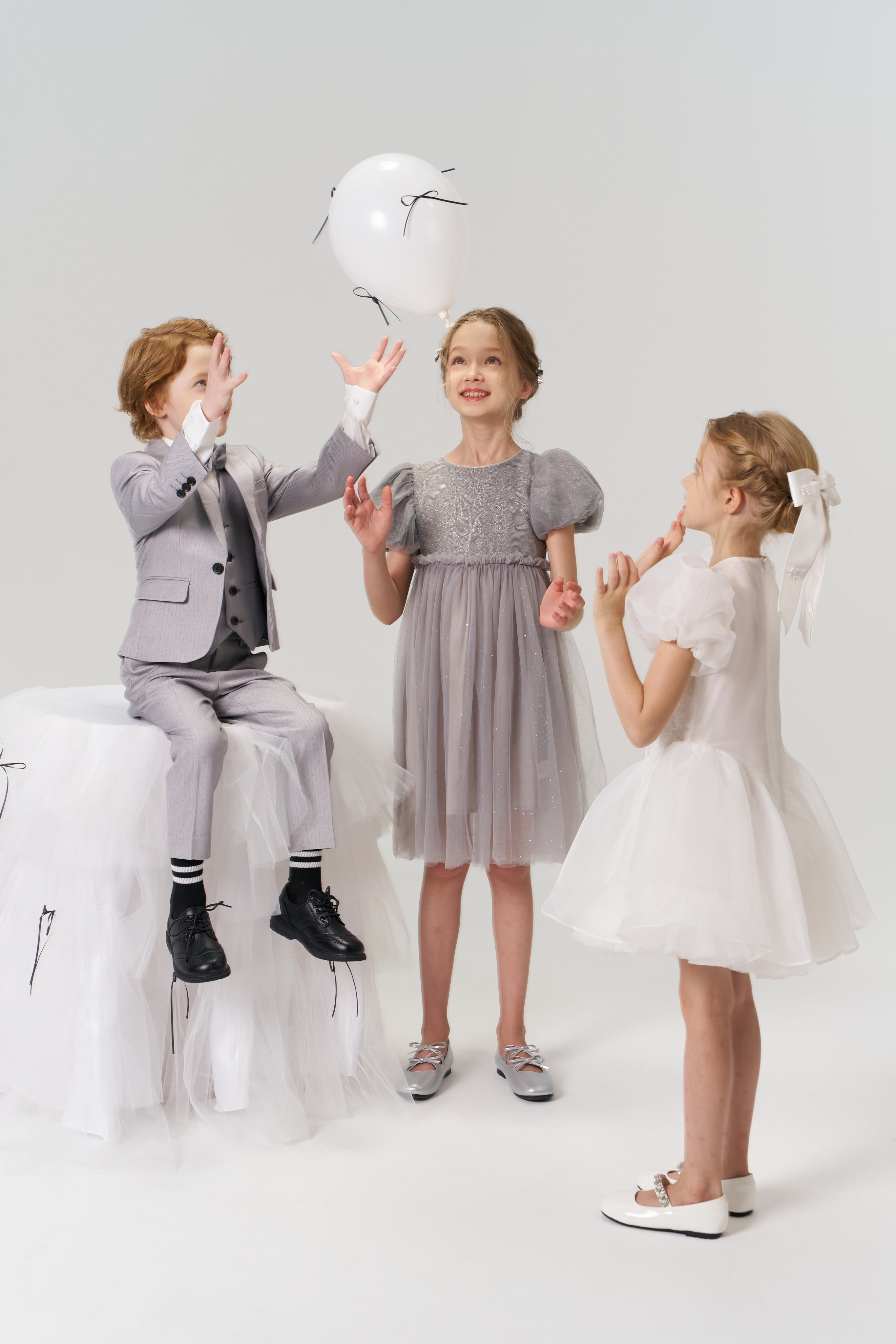 Three children play with a white balloon; one boy sits on a tulle stool while two girls stand nearby, with one wearing the Dusk Whisper Dress by KIKI and JOJO.