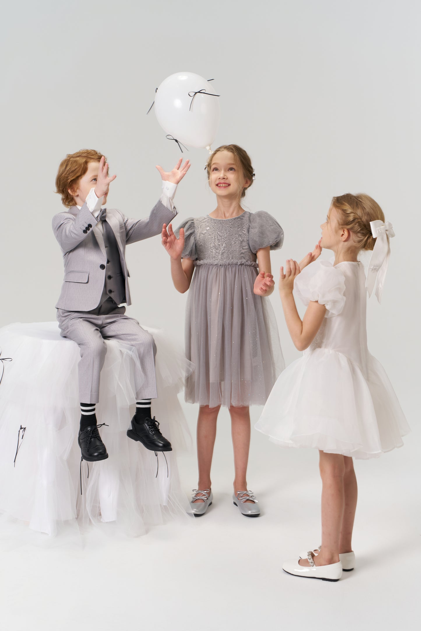 Three children play with a white balloon; one boy sits on a tulle stool while two girls stand nearby, with one wearing the Dusk Whisper Dress by KIKI and JOJO.