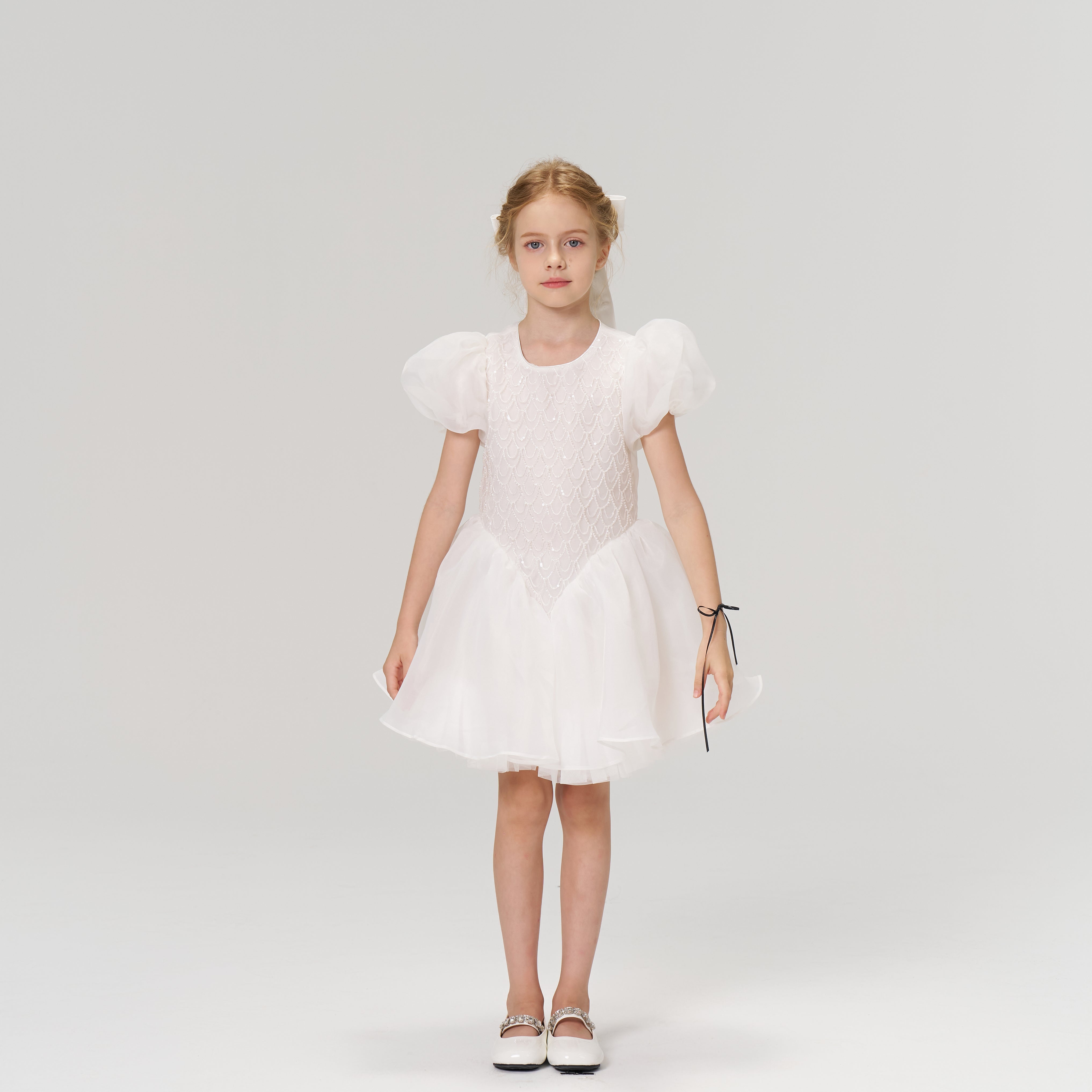 A young girl faces forward in the KIKI and JOJO Ballerina Whisper Dress—a white tulle mini dress with puffed sleeves—paired with white shoes, set against a plain light background.